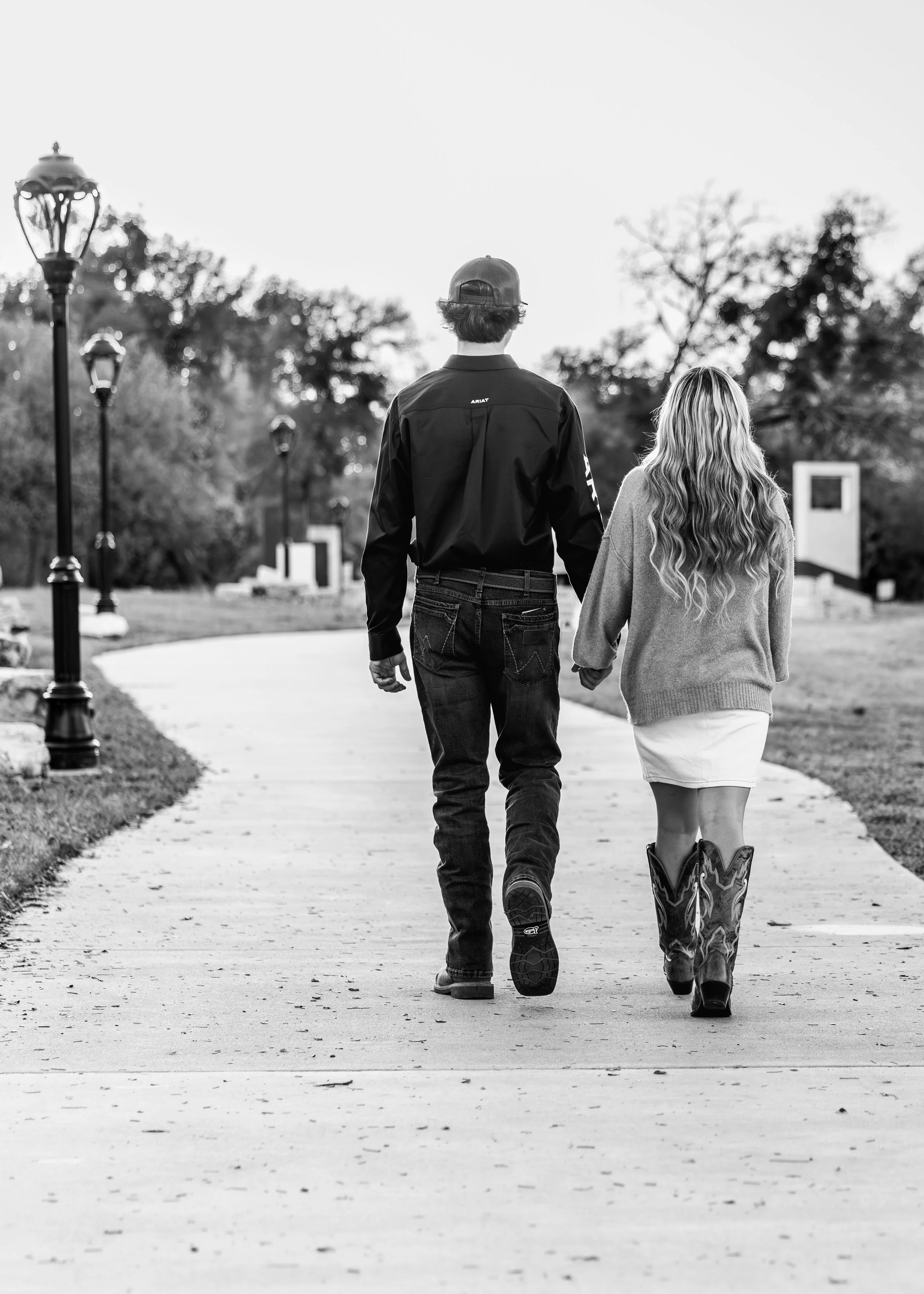 A black and white photo of a couple walking hand in hand on a paved path in a park, with trees and street lamps in the background.