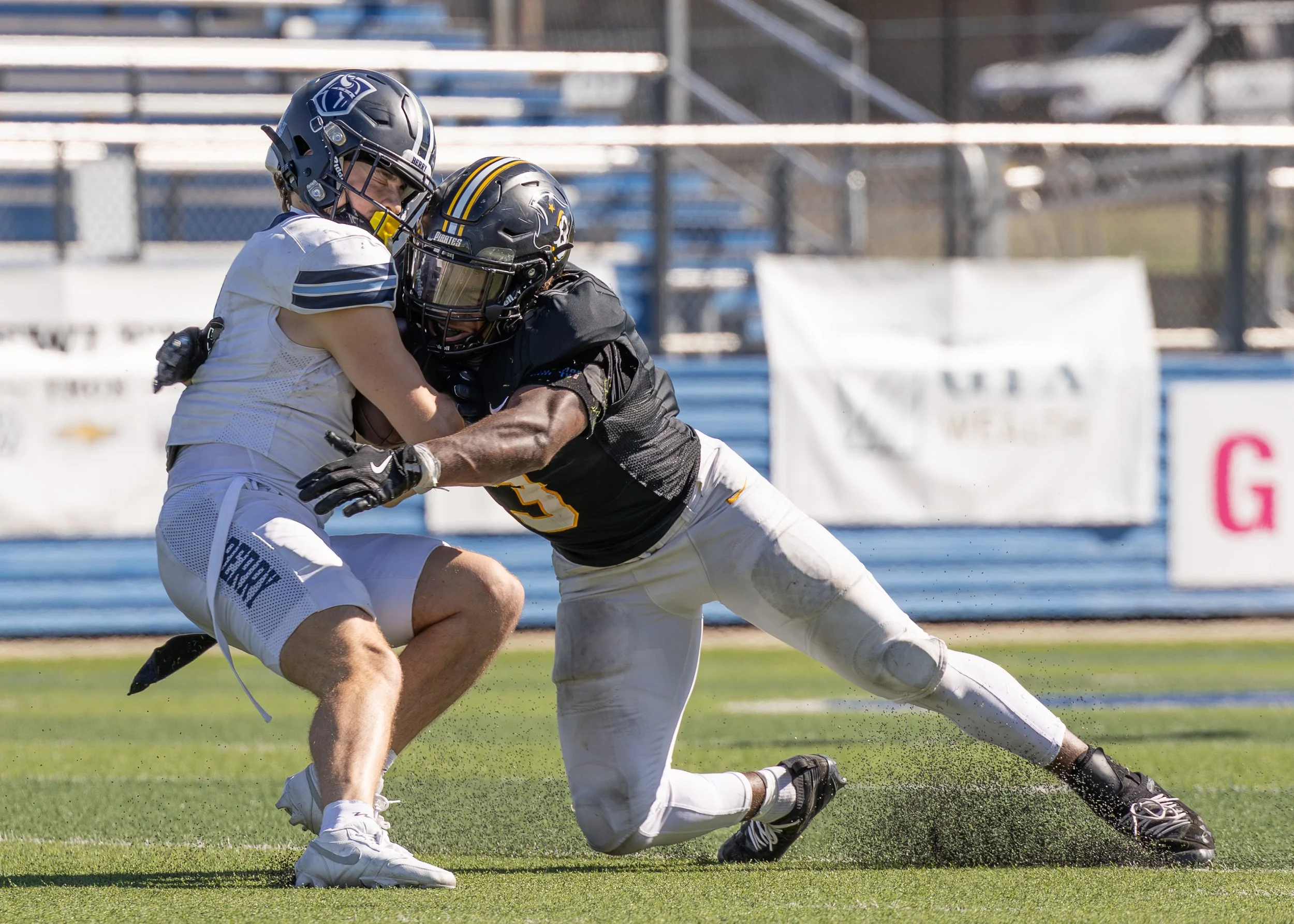 Two football players wearing helmets and uniforms are engaged in a tackle on a football field. One player in a white uniform is holding the football, while the other in a black uniform is attempting to bring him down.
