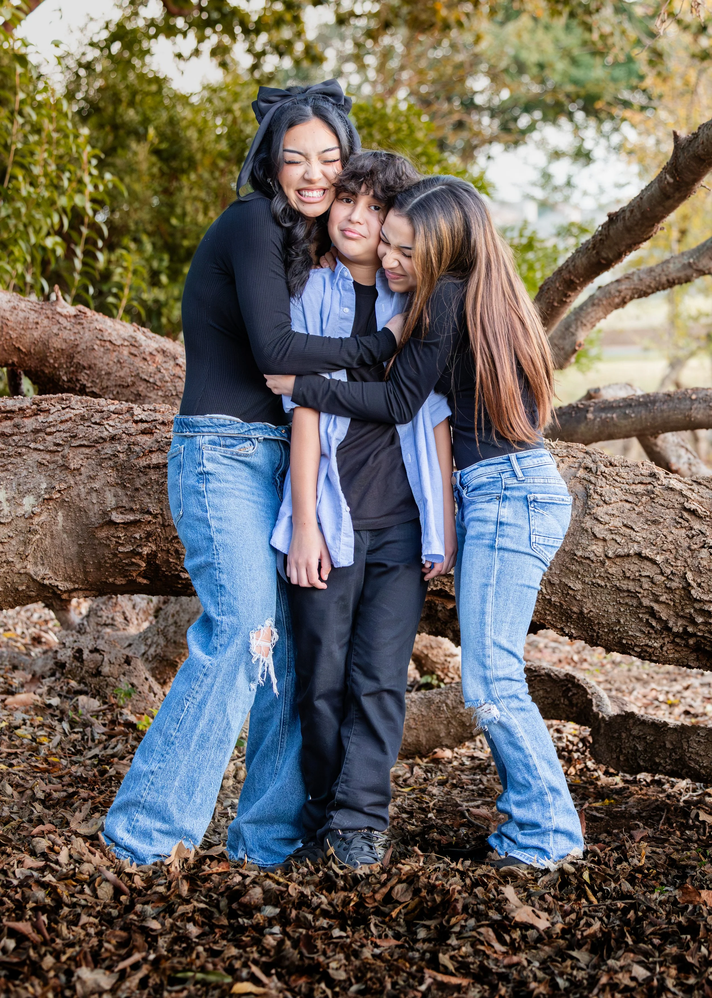 Three people hugging outdoors, standing on fallen leaves with a large tree trunk and branches behind them.