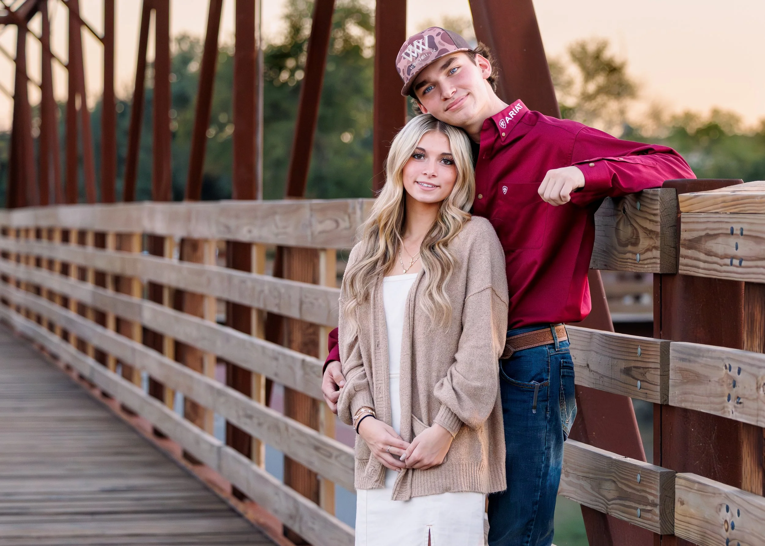 A young couple with light skin and blonde hair posing together on a wooden bridge during sunset, with trees in the background.