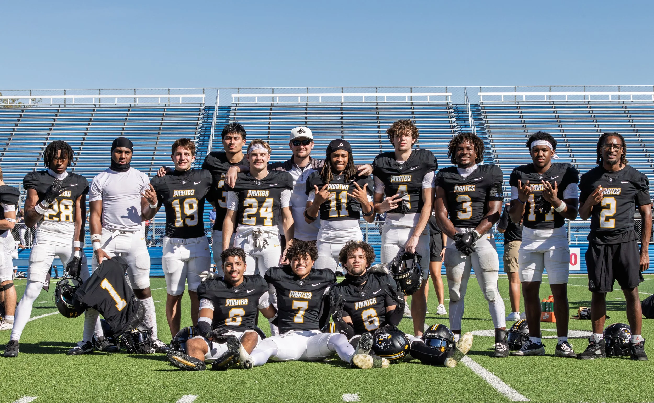 A football team in black and white uniforms posing on a football field, with some players kneeling in front and others standing behind, in front of empty bleachers.
