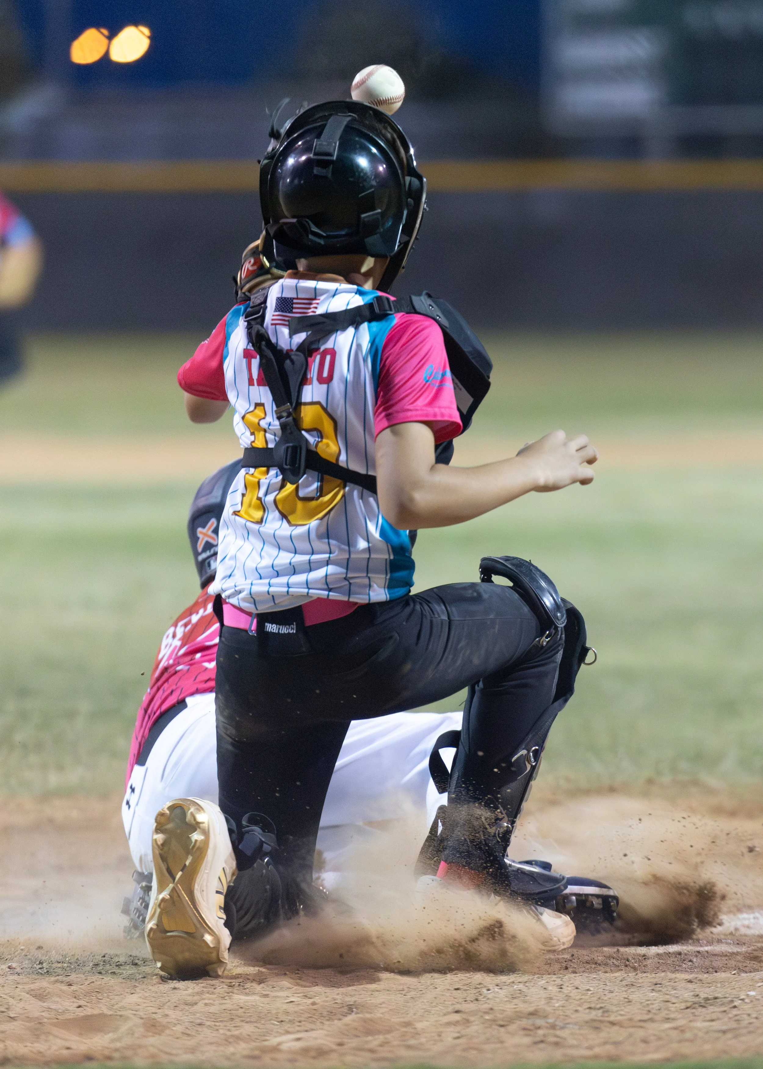 A baseball player, wearing a protective helmet and pink jersey, appears to have just been tagged out at home plate, while the catcher, dressed in red and white, is on the ground with a baseball in mid-air above them.