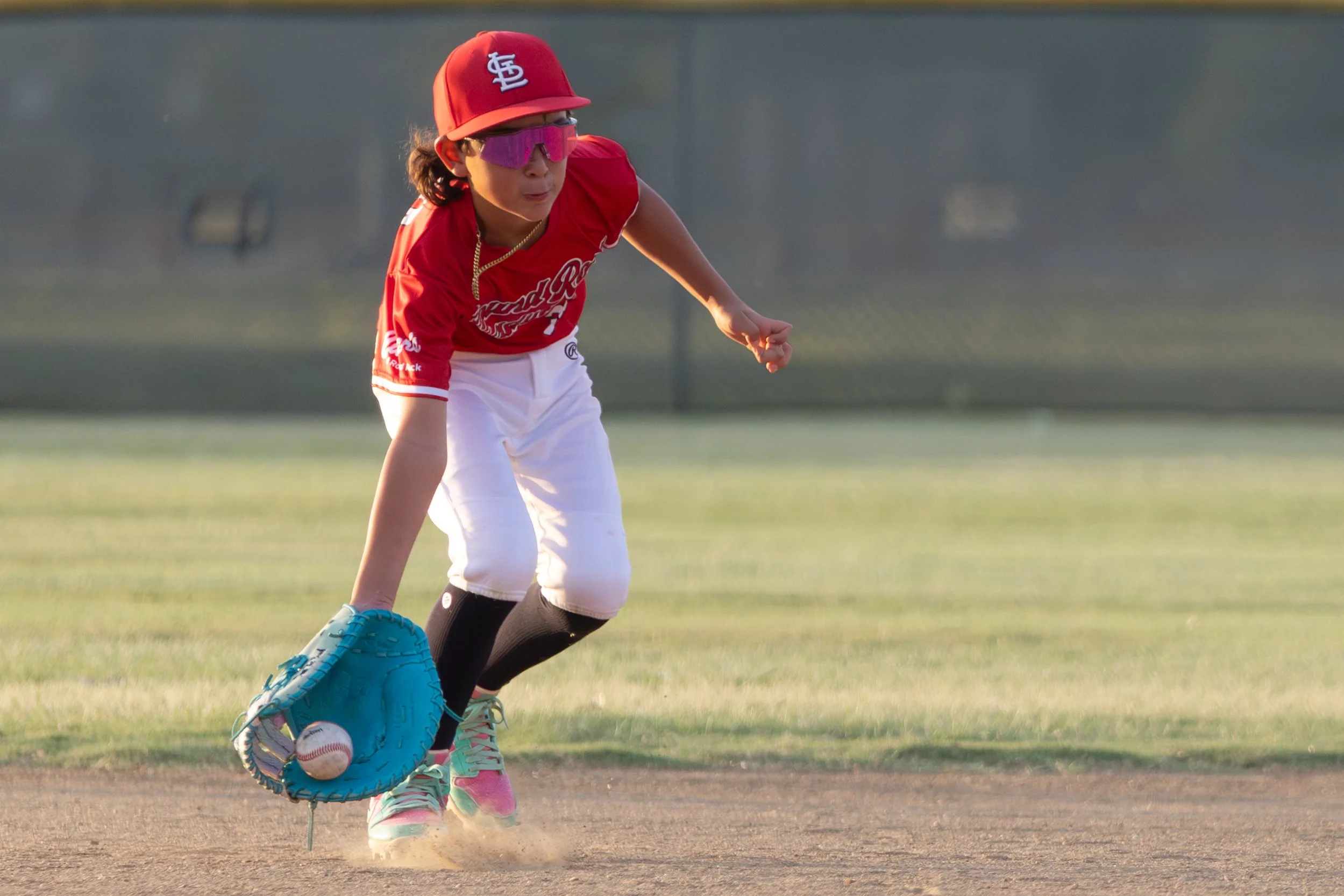 A young girl in baseball attire, wearing a red cap and jersey, white pants, and pink and green cleats, is on a baseball field fielding a ground ball with her glove.