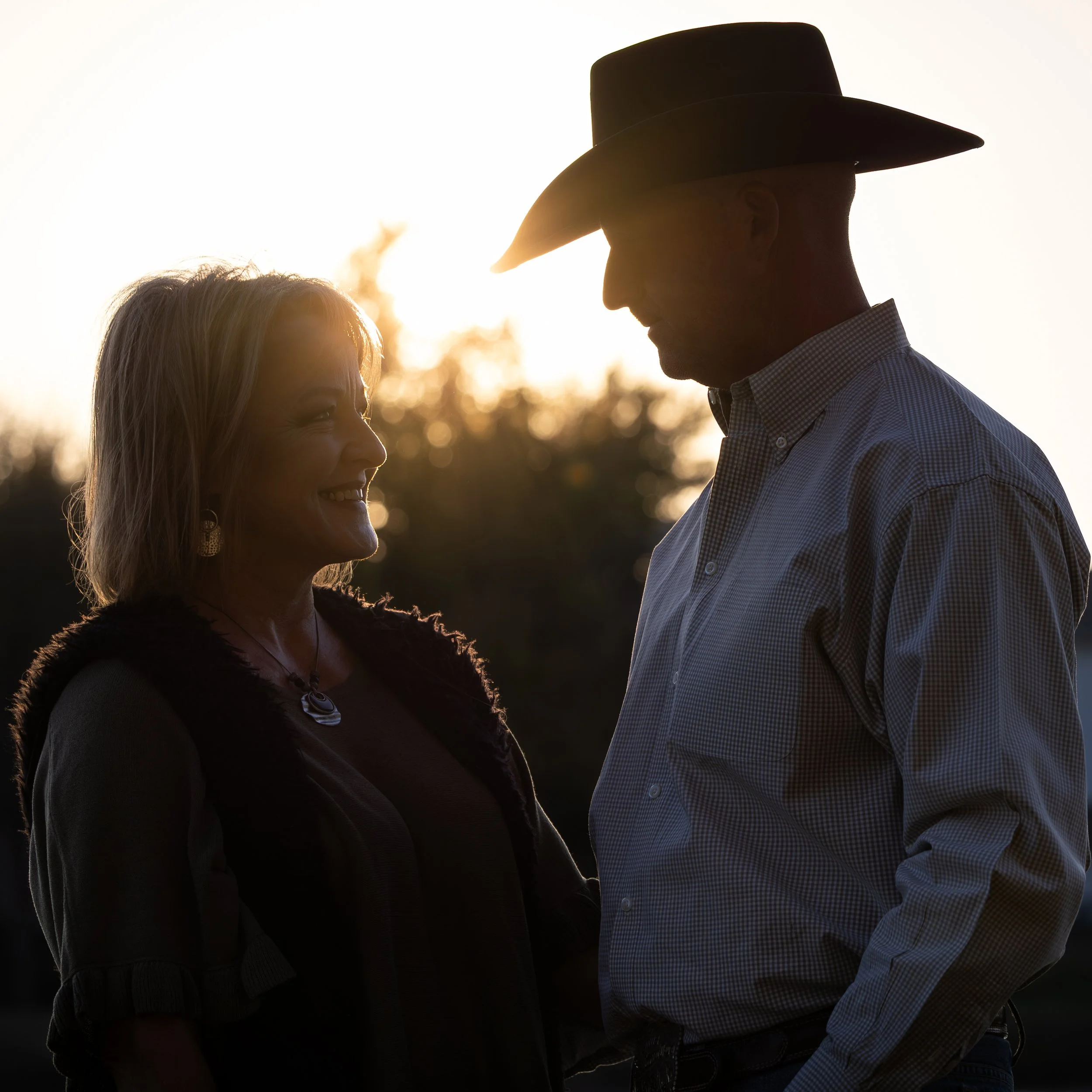 Silhouette of a man and woman facing each other outdoors at sunset, with the sun behind them creating a glowing background. The woman is smiling, and the man wears a cowboy hat.