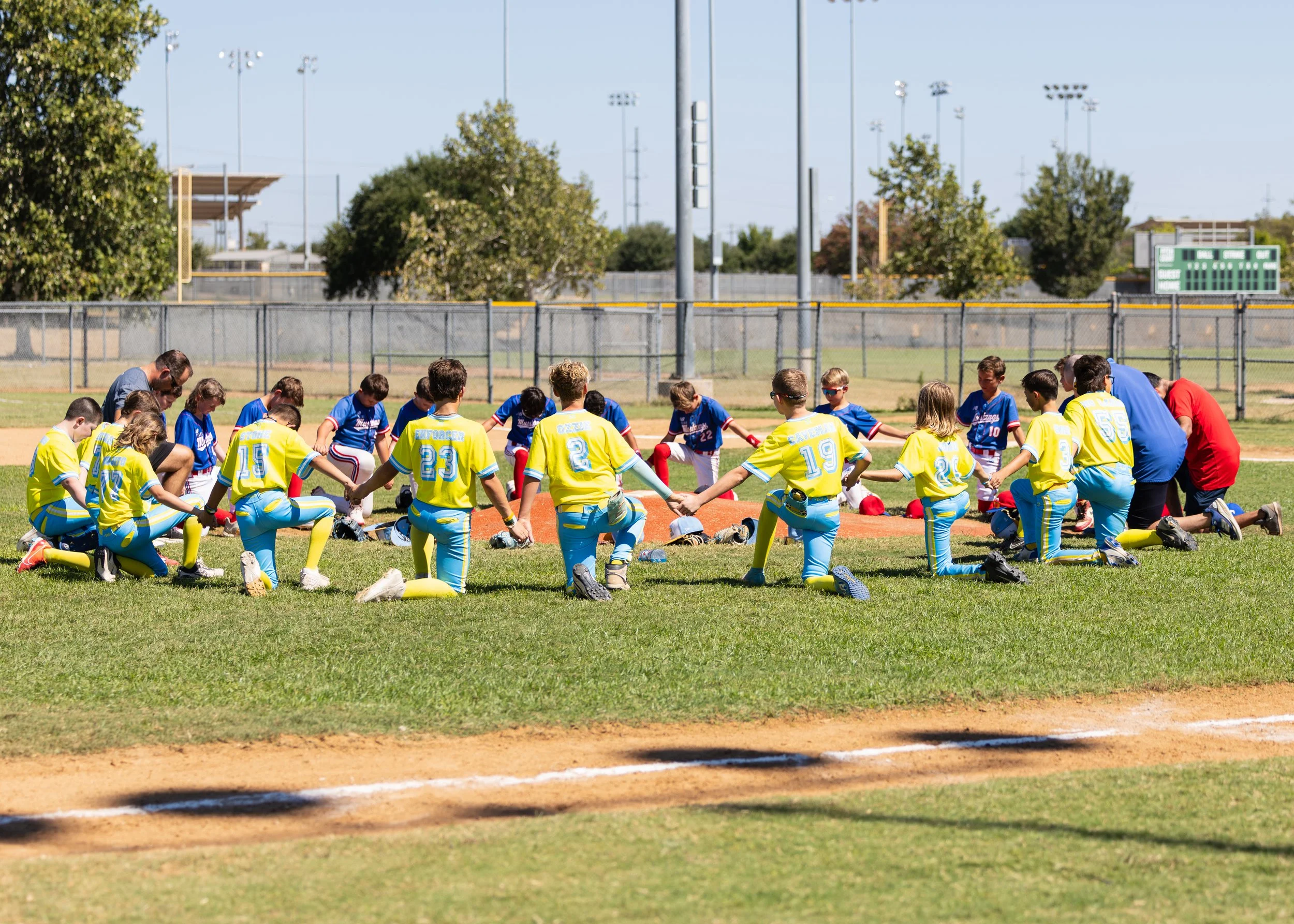 Youth baseball players kneeling in a prayer circle on the field before a game, with coaches and helmets in front of them.