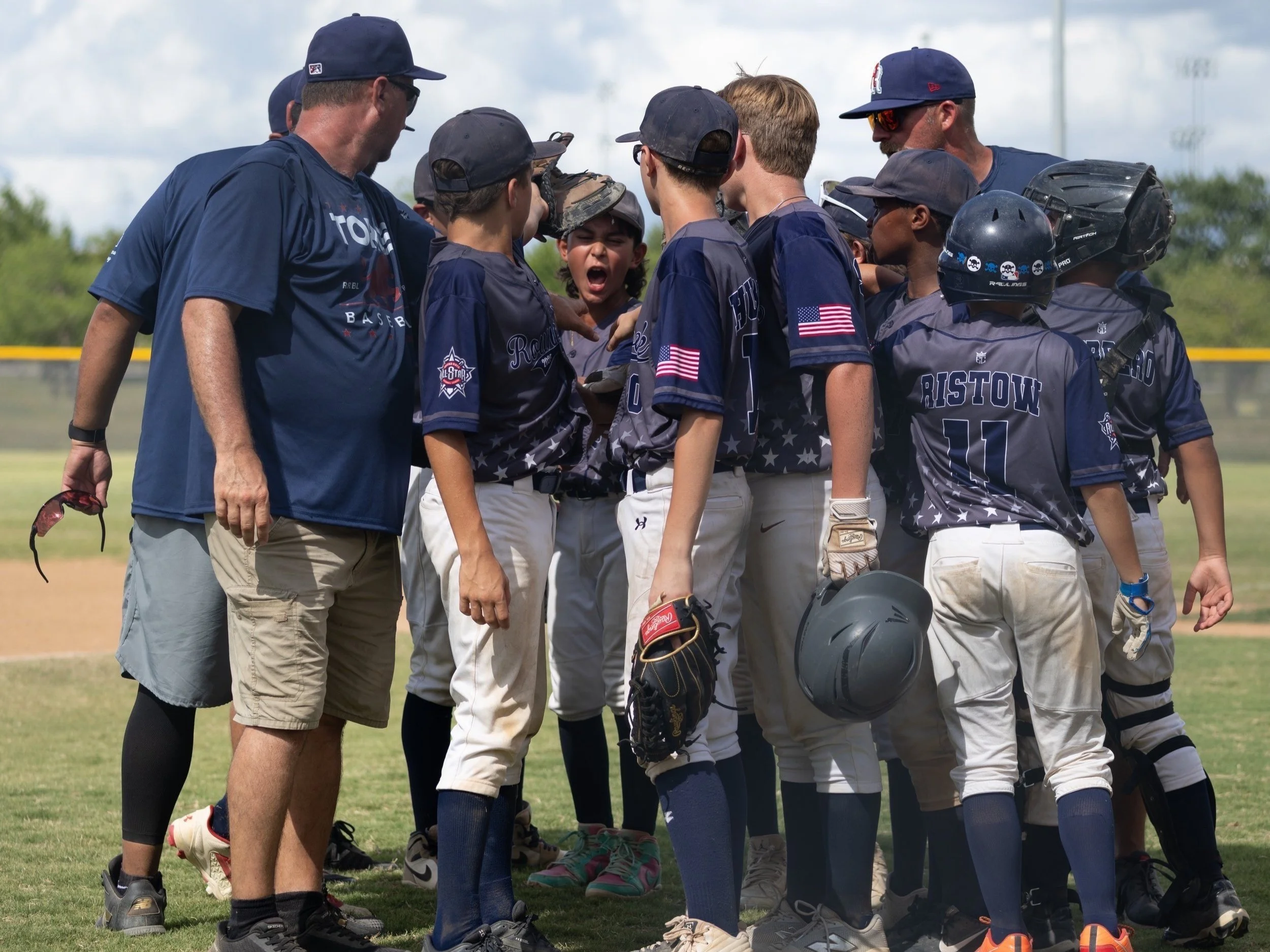 Youth baseball team gathered in a huddle with coach on baseball field during daytime.