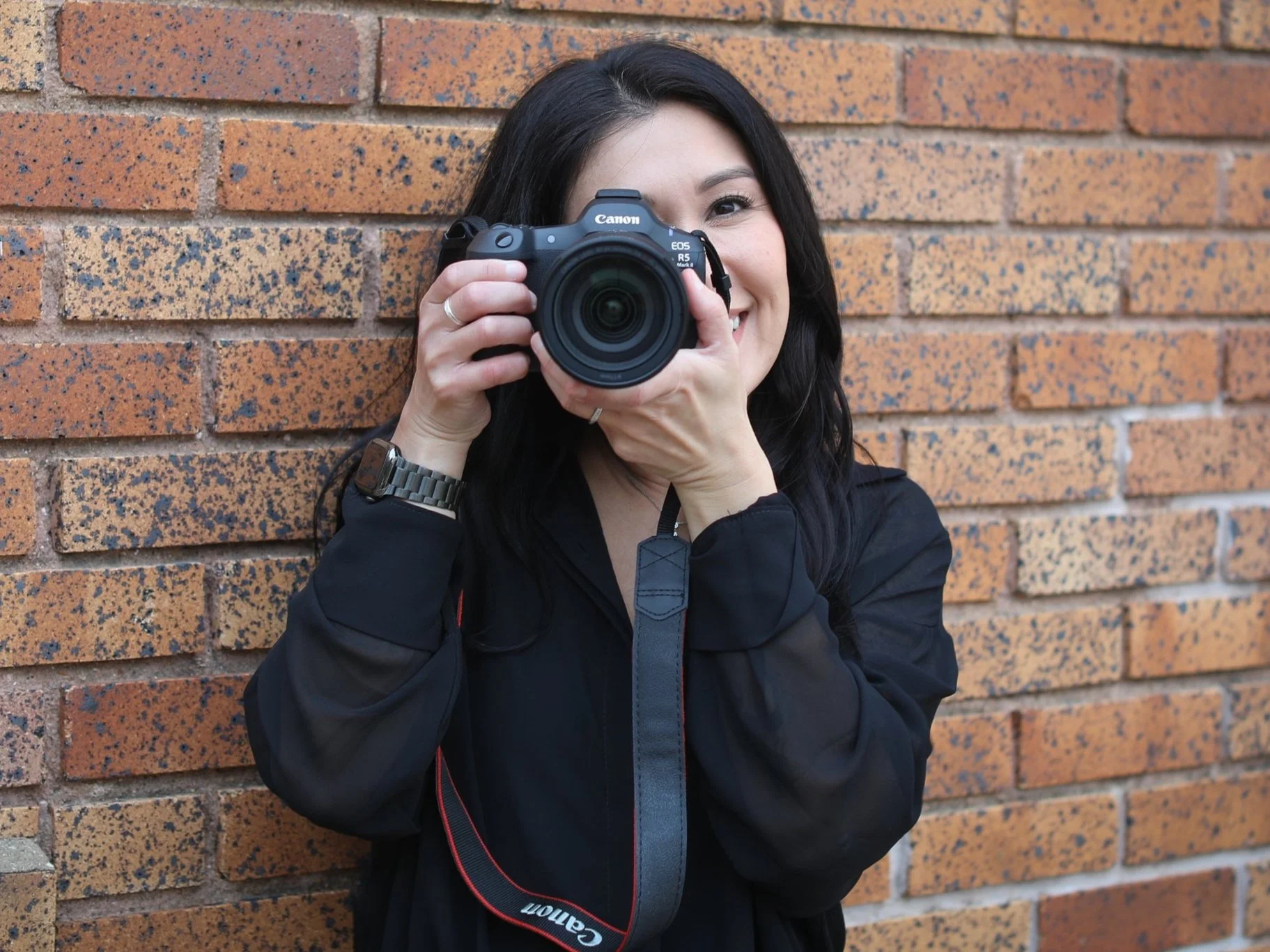 A woman with black hair taking a photo with a Canon DSLR camera against a brick wall.