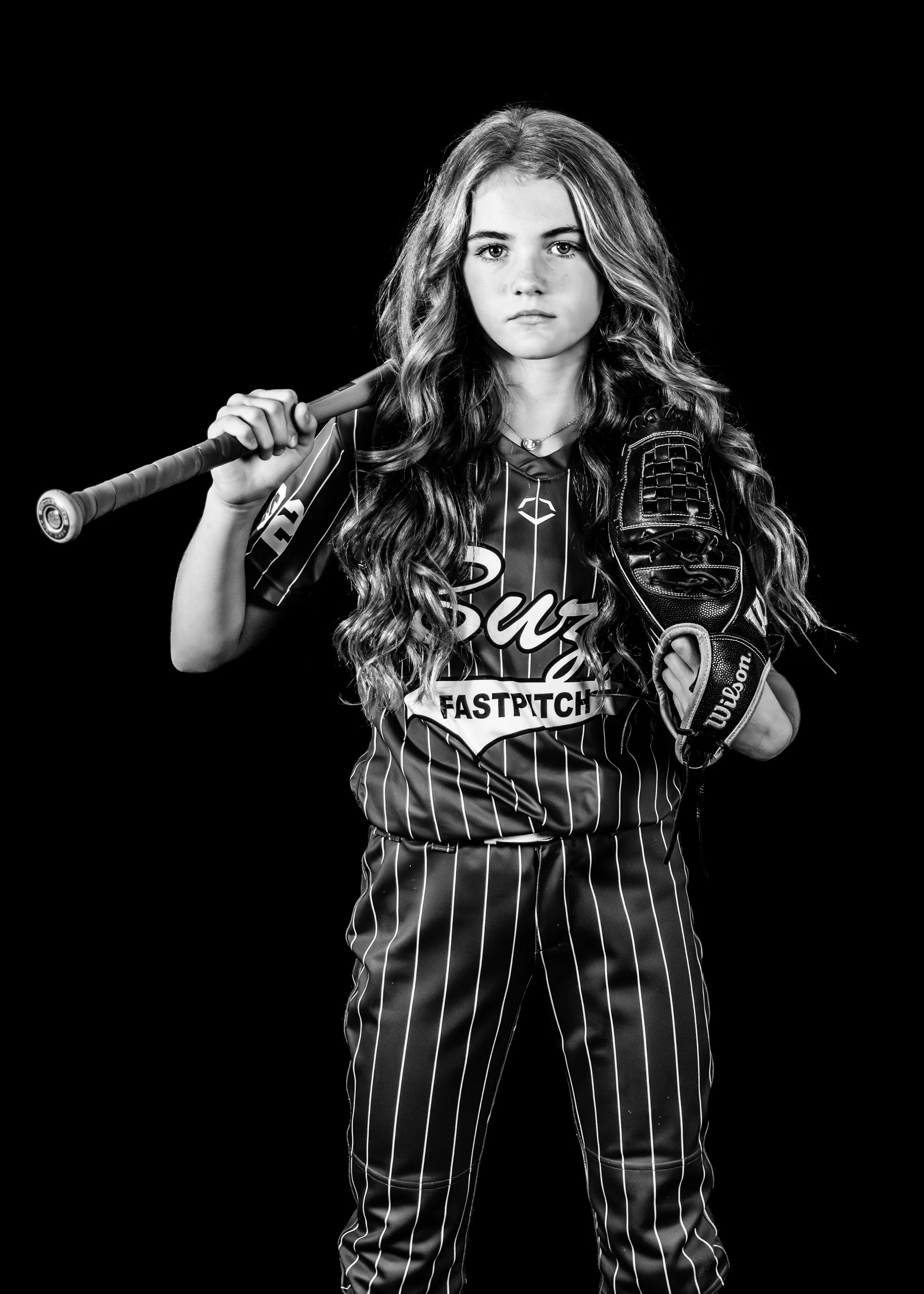 Black and white photo of a young girl in softball uniform holding a baseball bat over her shoulder and wearing a baseball glove on her left hand, standing against a black background.