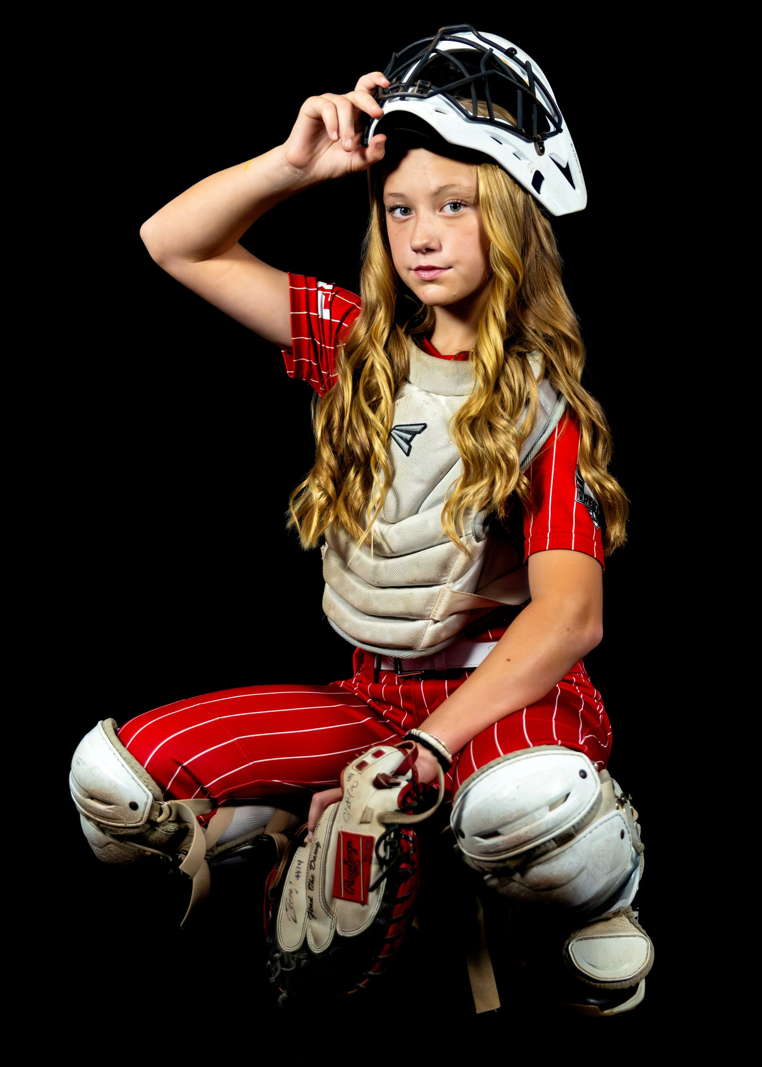 Young girl in baseball catcher's gear, holding helmet, sitting against black background.