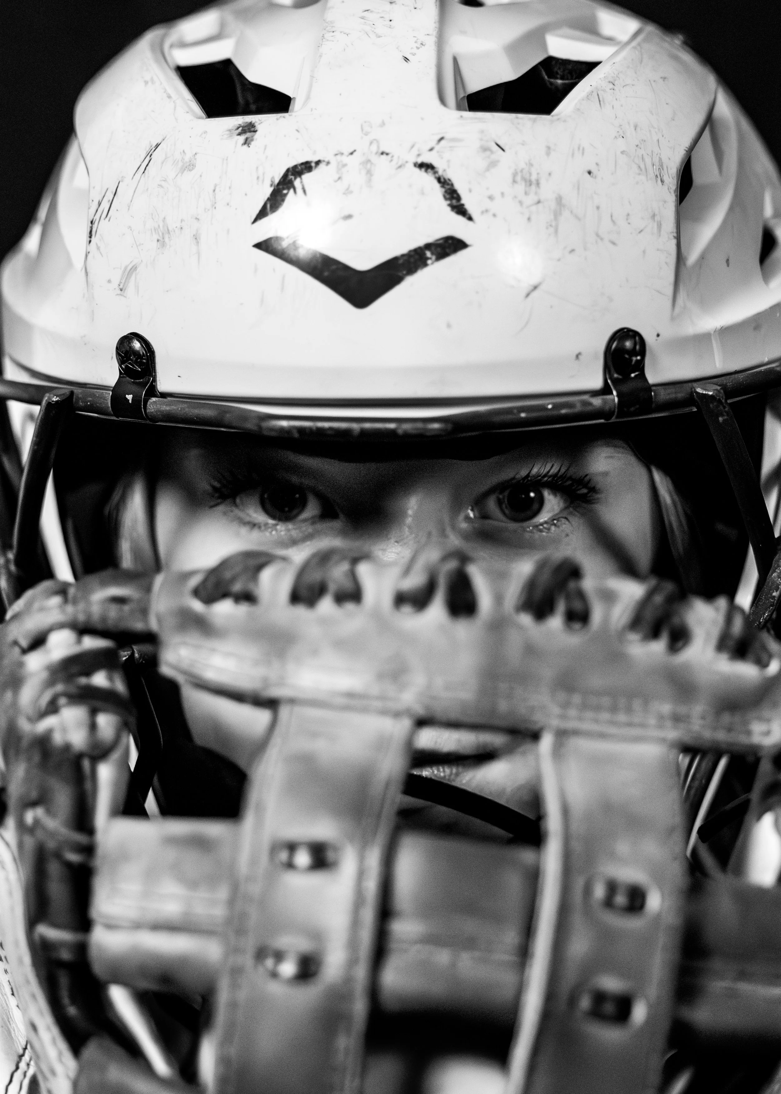 Close-up black-and-white photo of a young child's face wearing a sports helmet and holding a baseball glove near their face, with intense eyes visible.