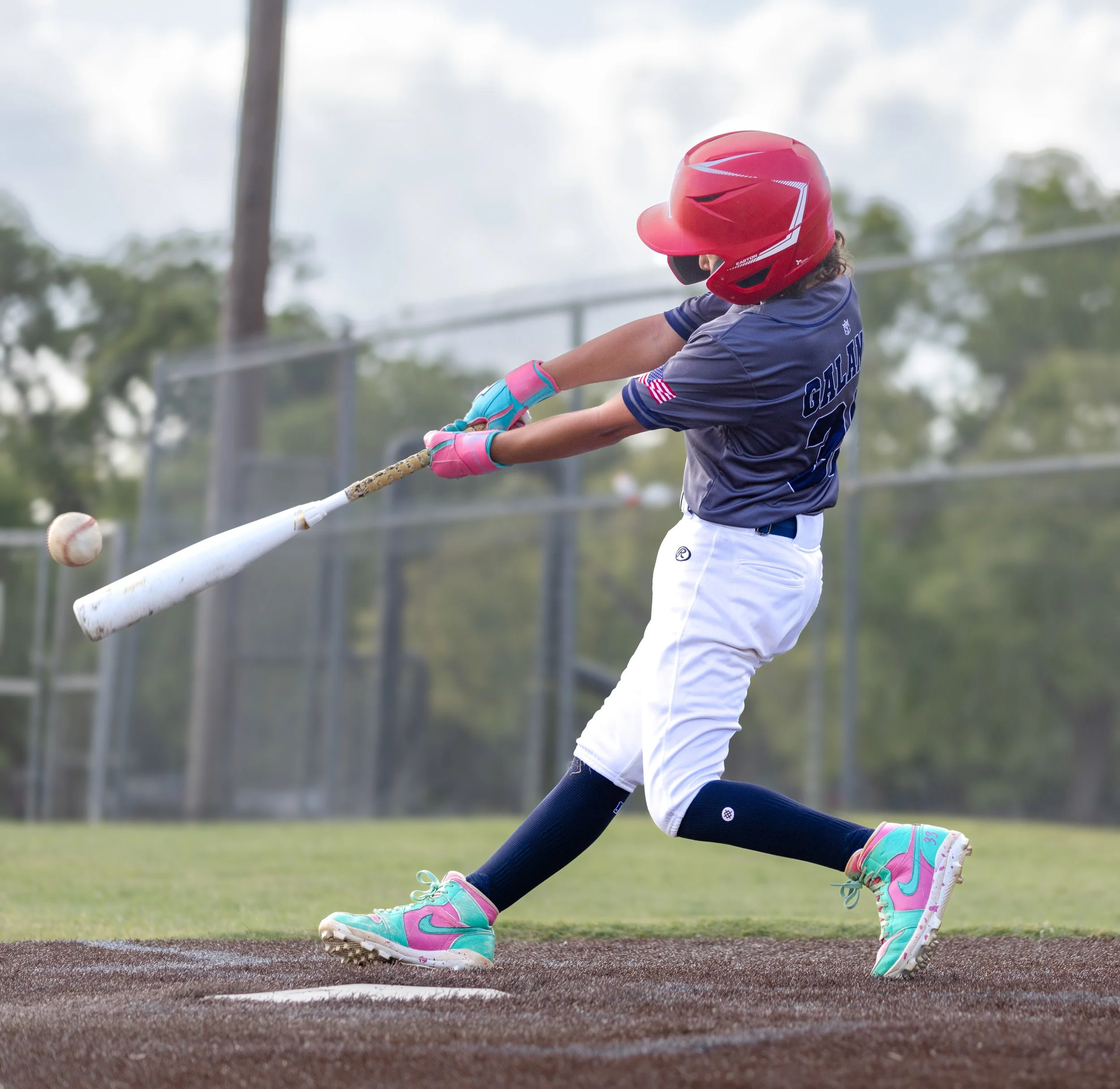 A young girl in a baseball uniform, wearing a red helmet and pink shoes, swings a bat at a baseball on a field.