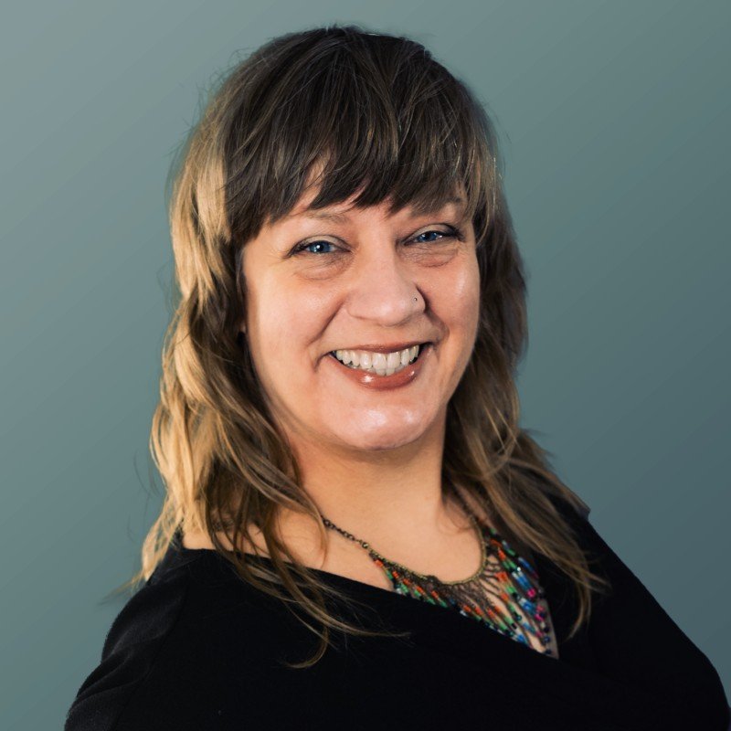 A woman with light brown, wavy hair, smiling, wearing a black top and a colorful beaded necklace, standing against a plain, muted green background.