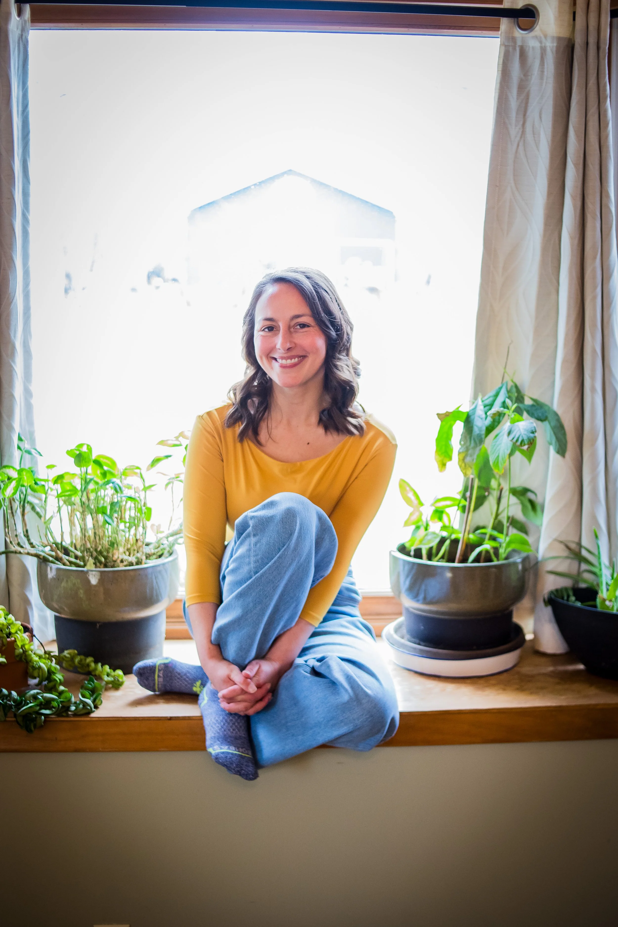 Jasmine, the founder of the Evolved Connection Project,  in yellow shirt sitting in window smiling with plants