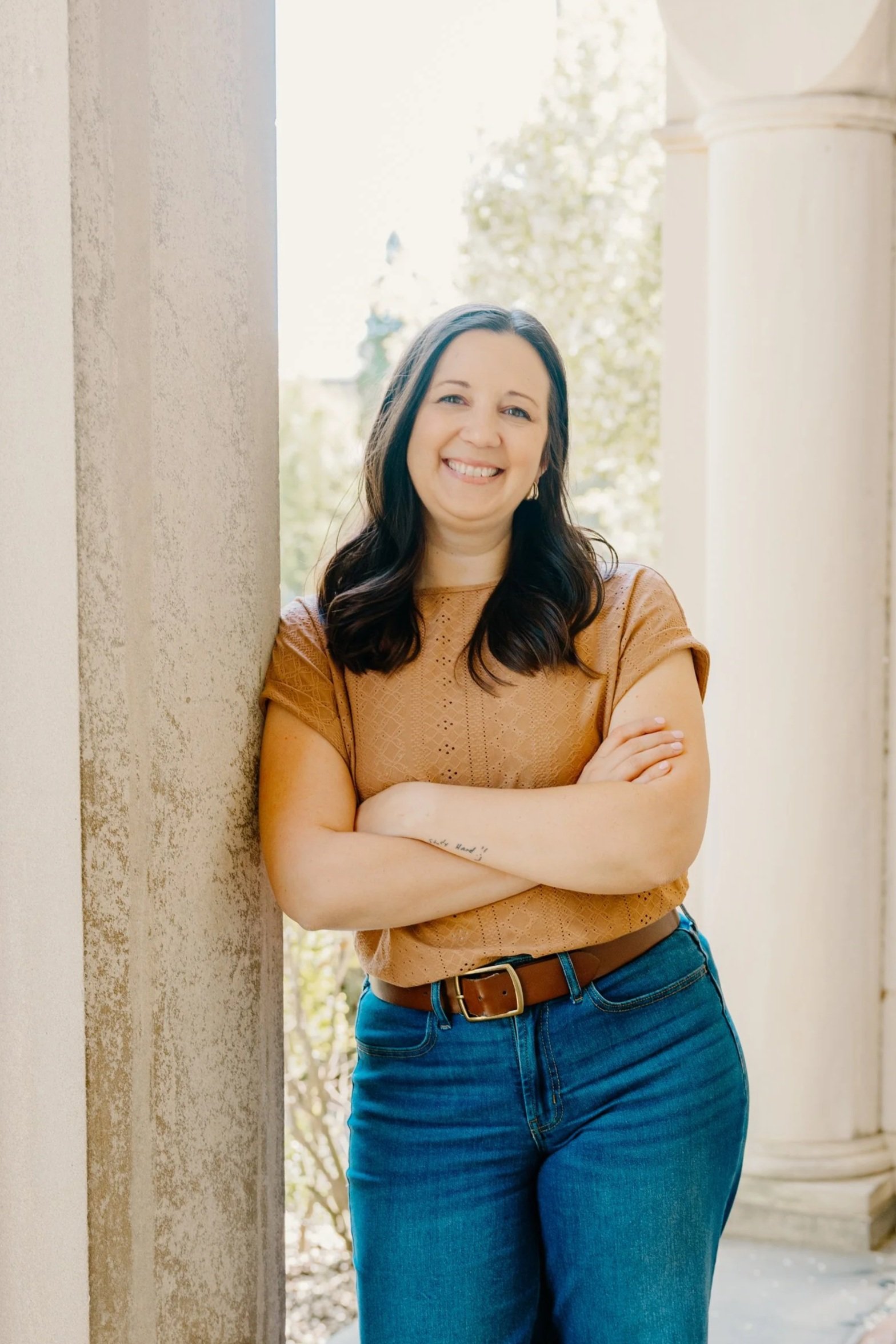 A woman with long dark hair wearing a tan top and blue jeans, standing outdoors and smiling with arms crossed, next to a light-colored wall with architectural columns in the background.