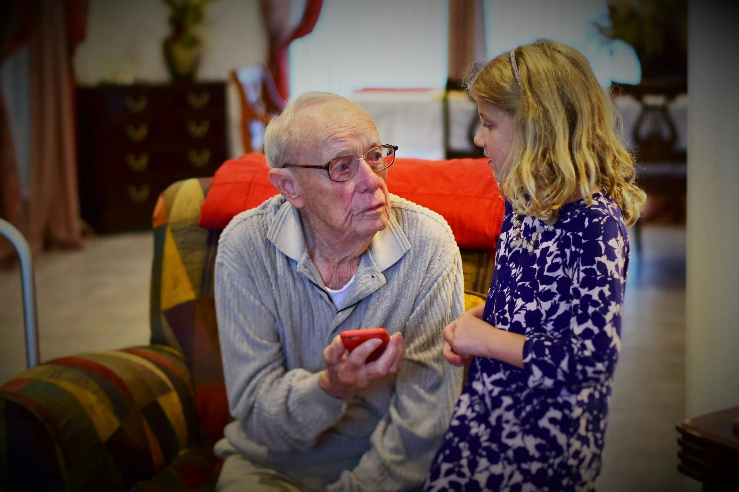 An elderly man with glasses holding a smartphone and talking with a young girl in a purple and white patterned dress in a cozy living room.