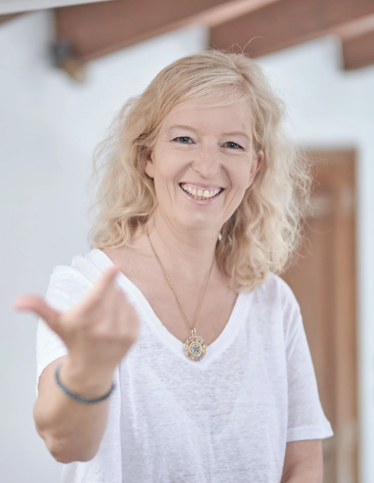 Eine lachende Frau mit blonden, lockigen Haaren trägt ein weißes T-Shirt und eine goldene Halskette, zeigt mit der Hand auf die Kamera.
