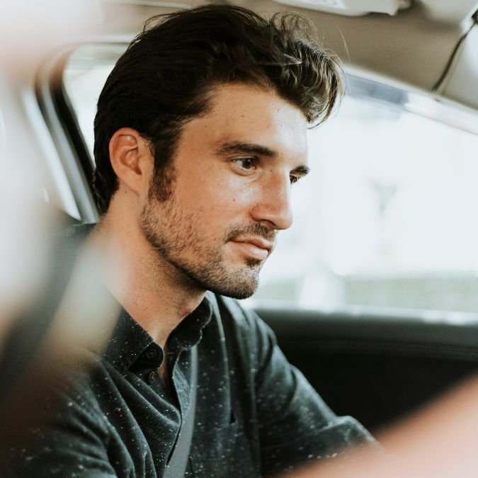 Ein junger Mann sitzt im Auto, schaut nachdenklich und trägt ein schwarzes Hemd.