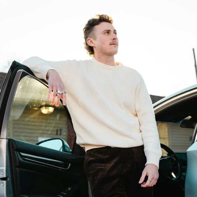 Junge Mann lehnt an einem Auto, trägt weißen Pullover und schwarze Hose, steht draußen.