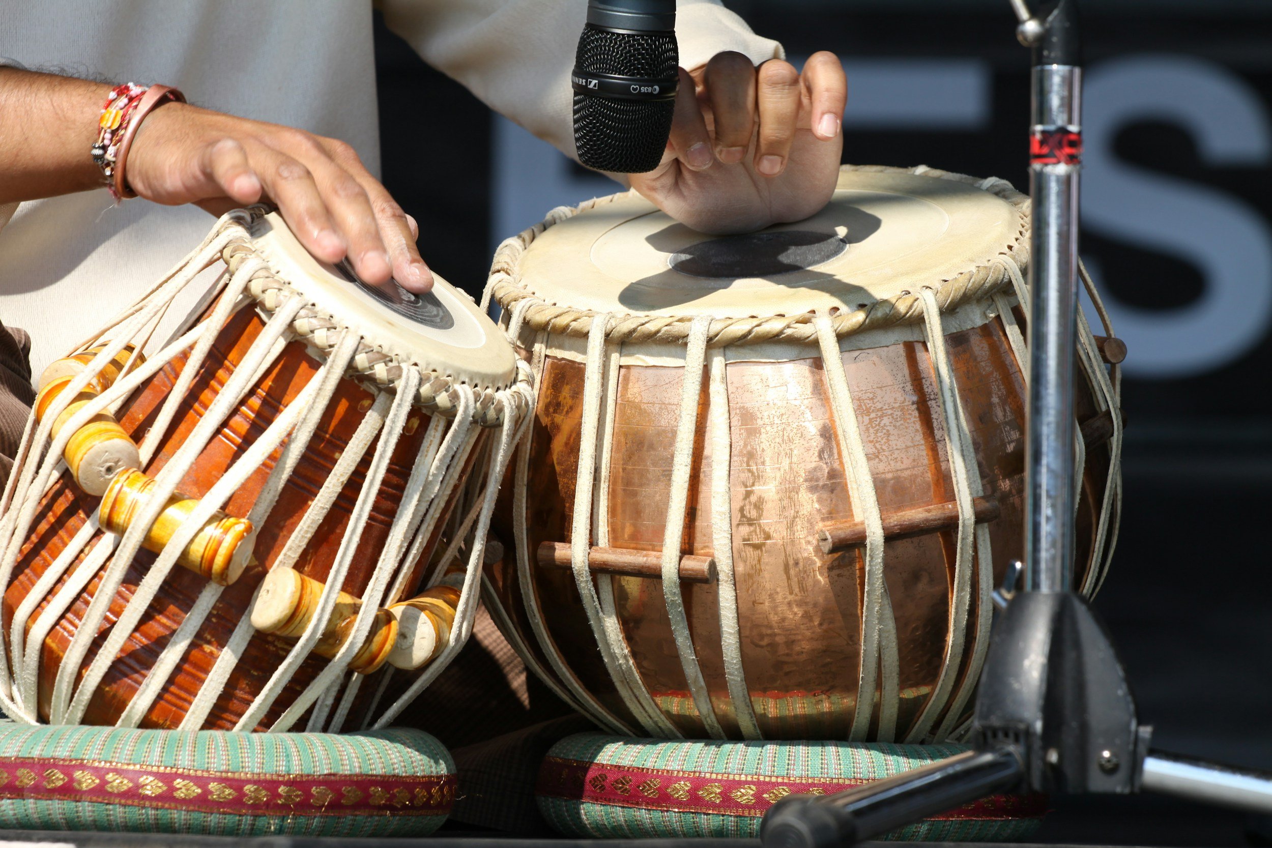 Two hands playing traditional Indian drums called tabla, one hand is striking the smaller drum, with a microphone positioned nearby.