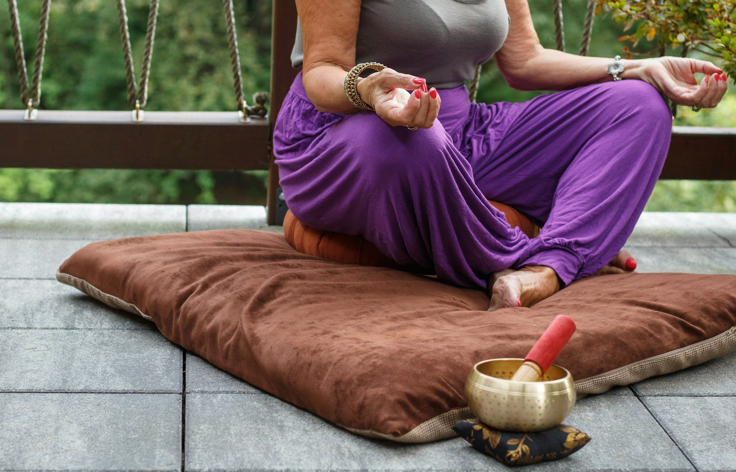 A person sitting cross-legged on an outdoor cushion, practicing meditation with a singing bowl and mallet nearby.