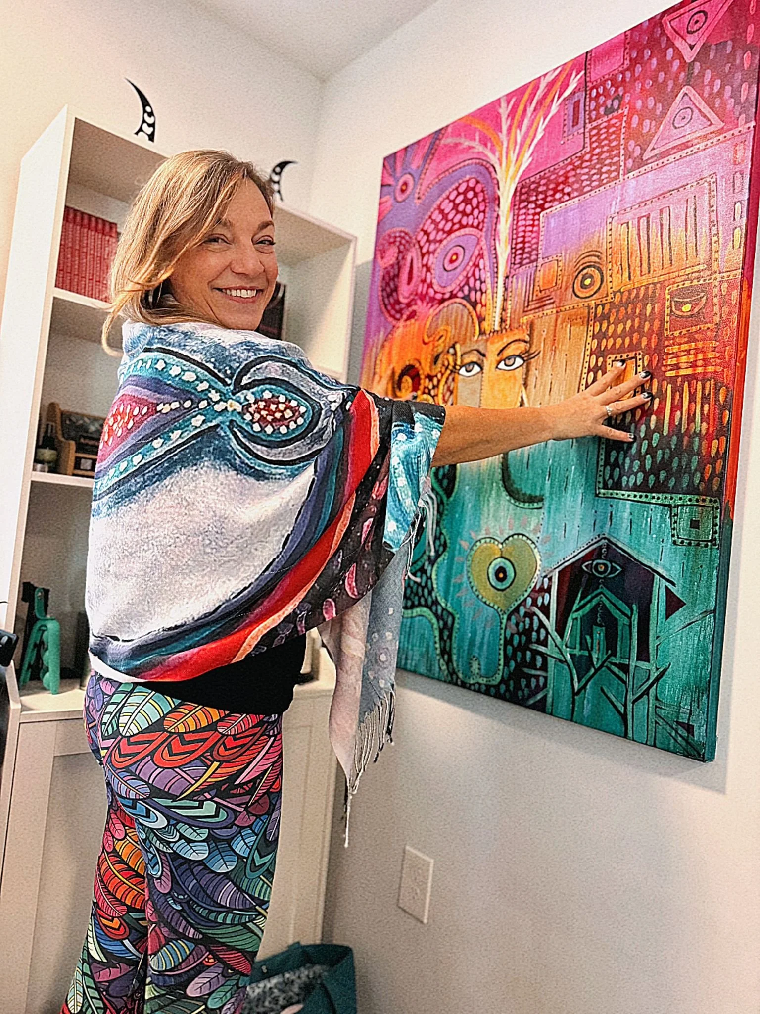 Woman smiling and pointing at a colorful abstract painting on a wall, dressed in vibrant, patterned clothing.