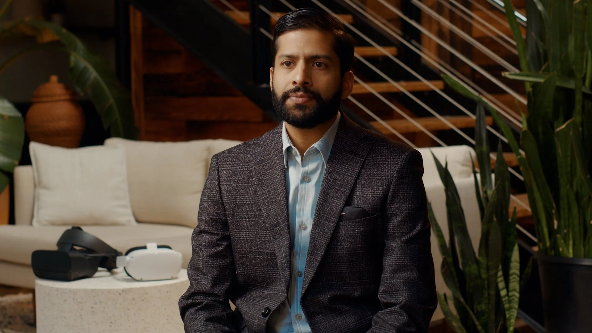 A man with dark hair and a beard, wearing a gray plaid suit and light blue shirt, sitting in a modern living room with green plants and a white sofa, looking slightly to the side.