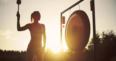 A woman silhouetted at sunset striking a gong with a mallet.