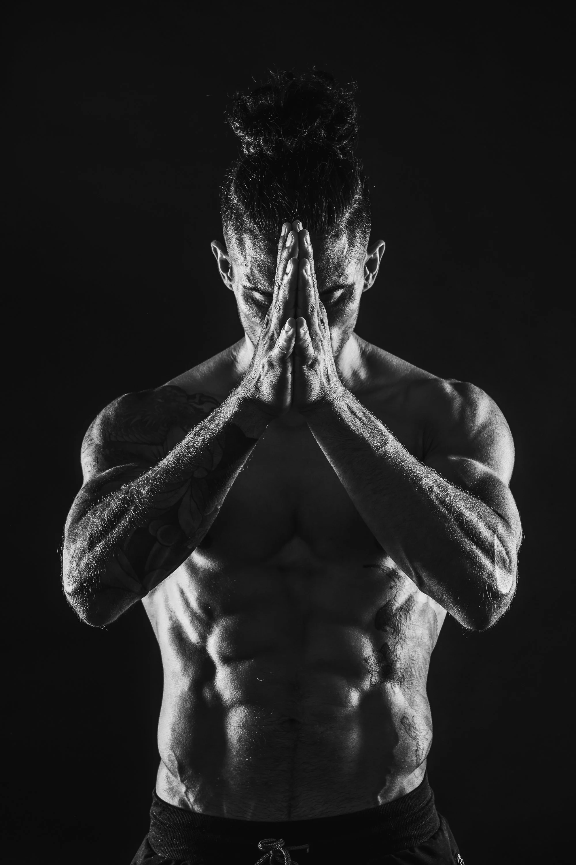 Black and white photo of a shirtless muscular man with tattoos, standing against a dark background, with his hands pressed together in a prayer pose and head bowed.