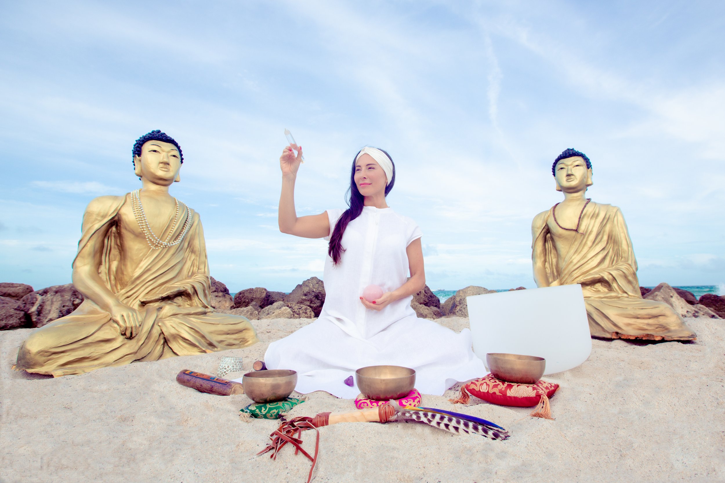 A woman dressed in white robes and headscarf sitting on sandy beach with her eyes closed, holding a pink crystal ball in her left hand and a small white object in her right hand, surrounded by spiritual items including bowls, feathers, and sculptures of seated Buddha figures.