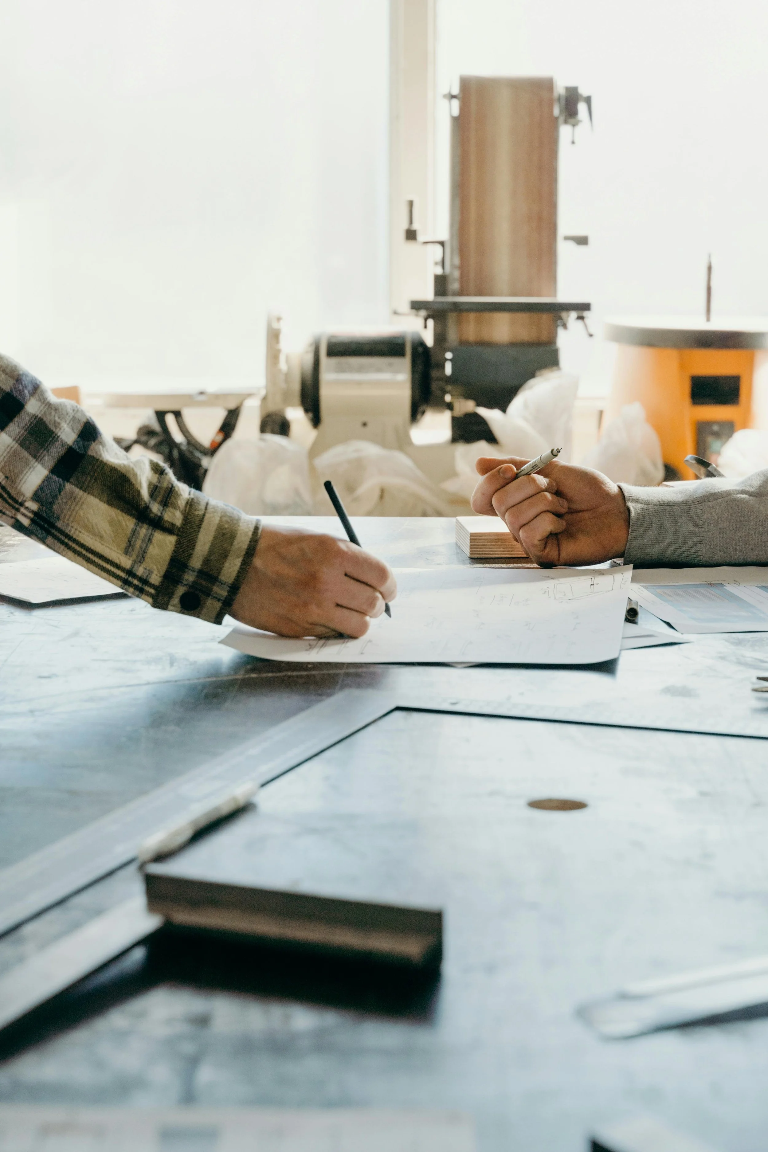 Two people working at a desk, one in a plaid shirt and the other in a gray hoodie, with drawings and documents on the table, in a workshop or studio setting.