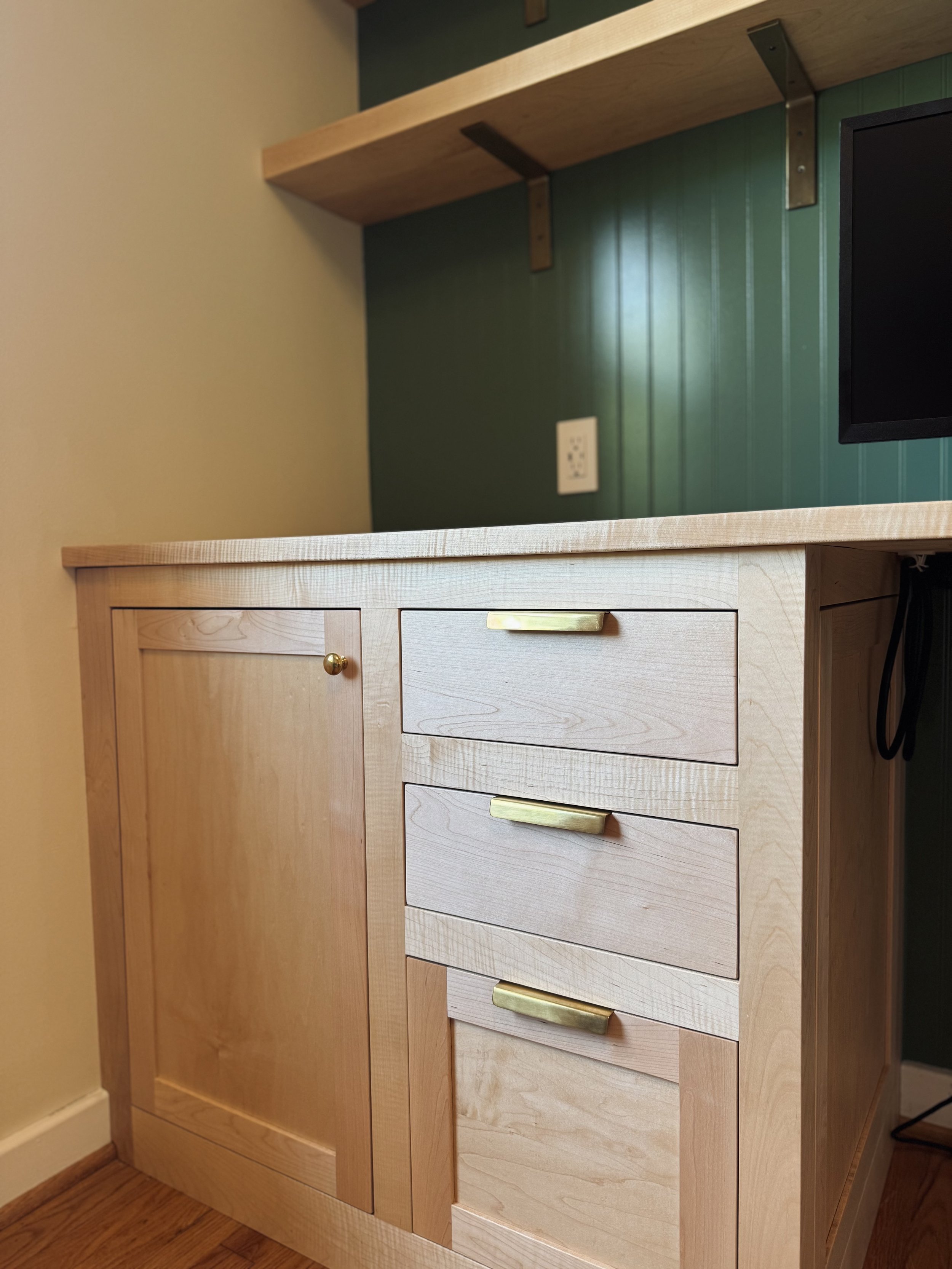 Close-up of a light wood cabinet with brass handles, featuring a large door and three drawers, in a room with a green wall and a wooden shelf above.