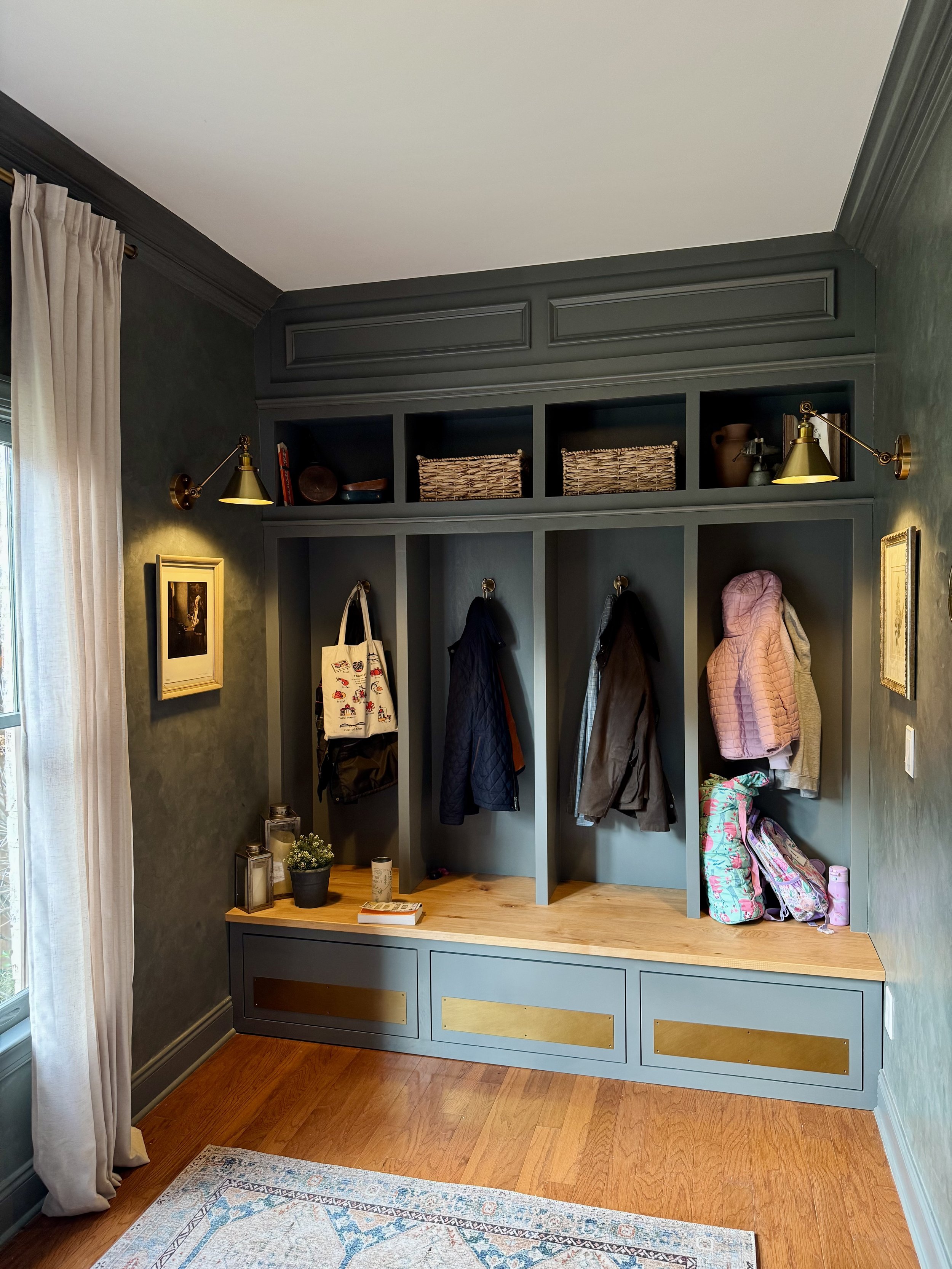 A built-in wall mudroom with blue cabinetry, hanging hooks with coats, and a wooden bench, decorated with baskets and lamps.