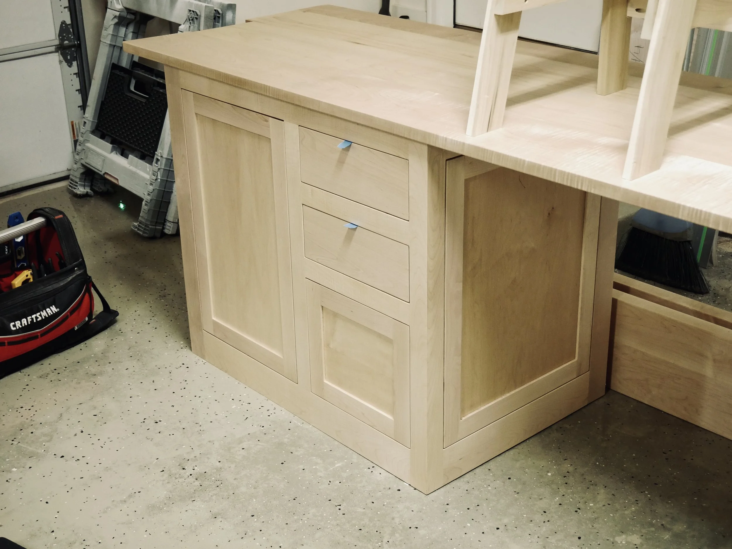 Unfinished wooden kitchen cabinet with three drawers and a door, situated in a workshop with tools and a ladder nearby.