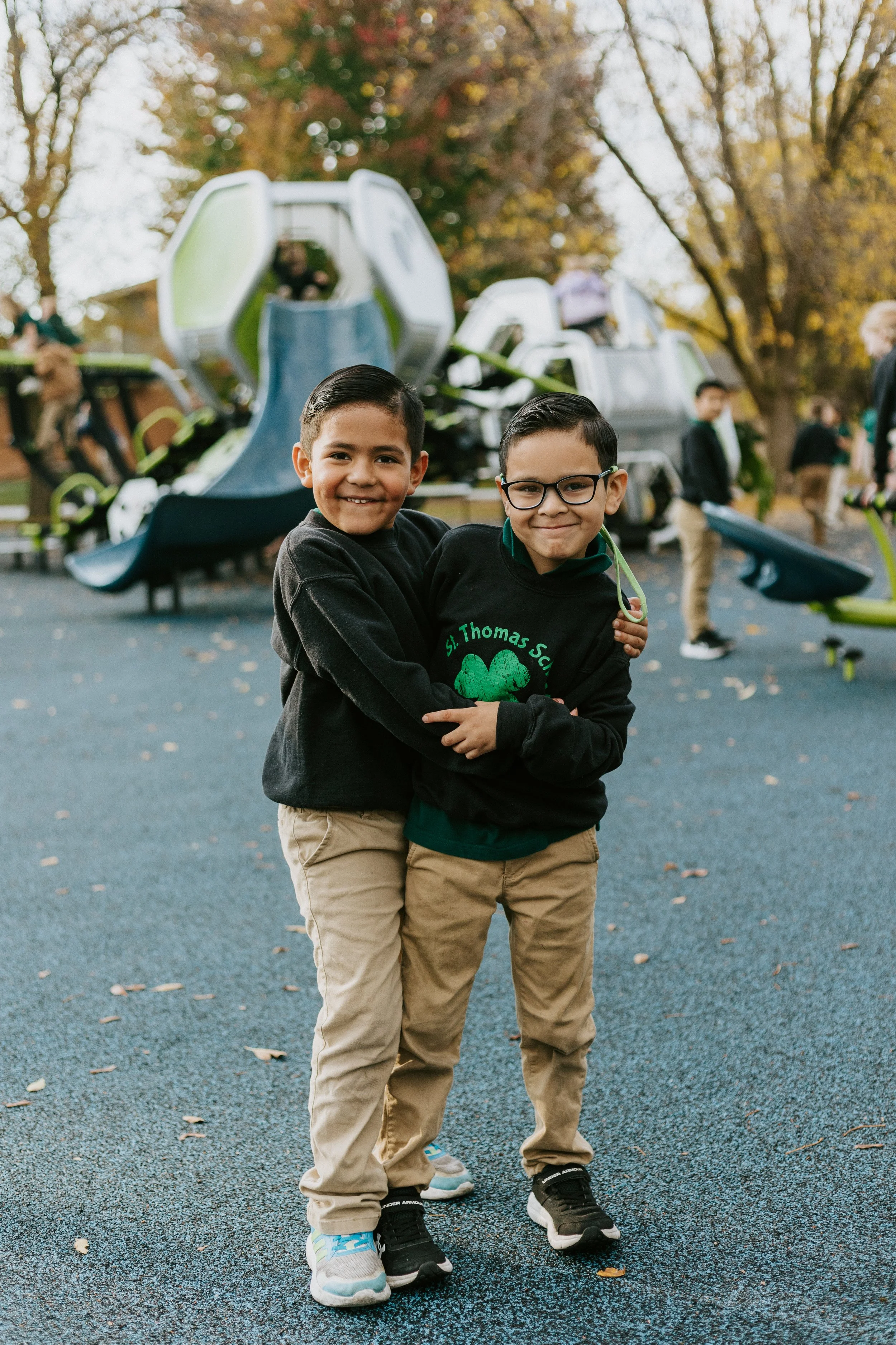 Two young boys hugging and smiling on a playground, with a modern play structure in the background, surrounded by trees with autumn leaves.
