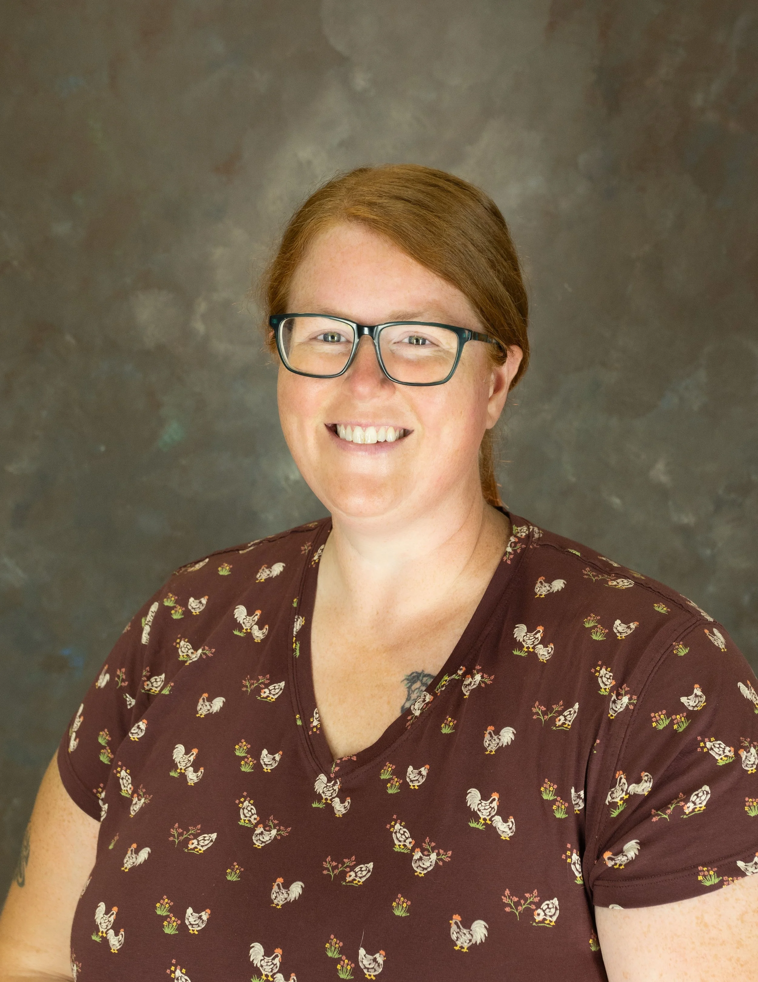 A woman with red hair, glasses, and freckles, smiling at the camera against a dark, textured background.