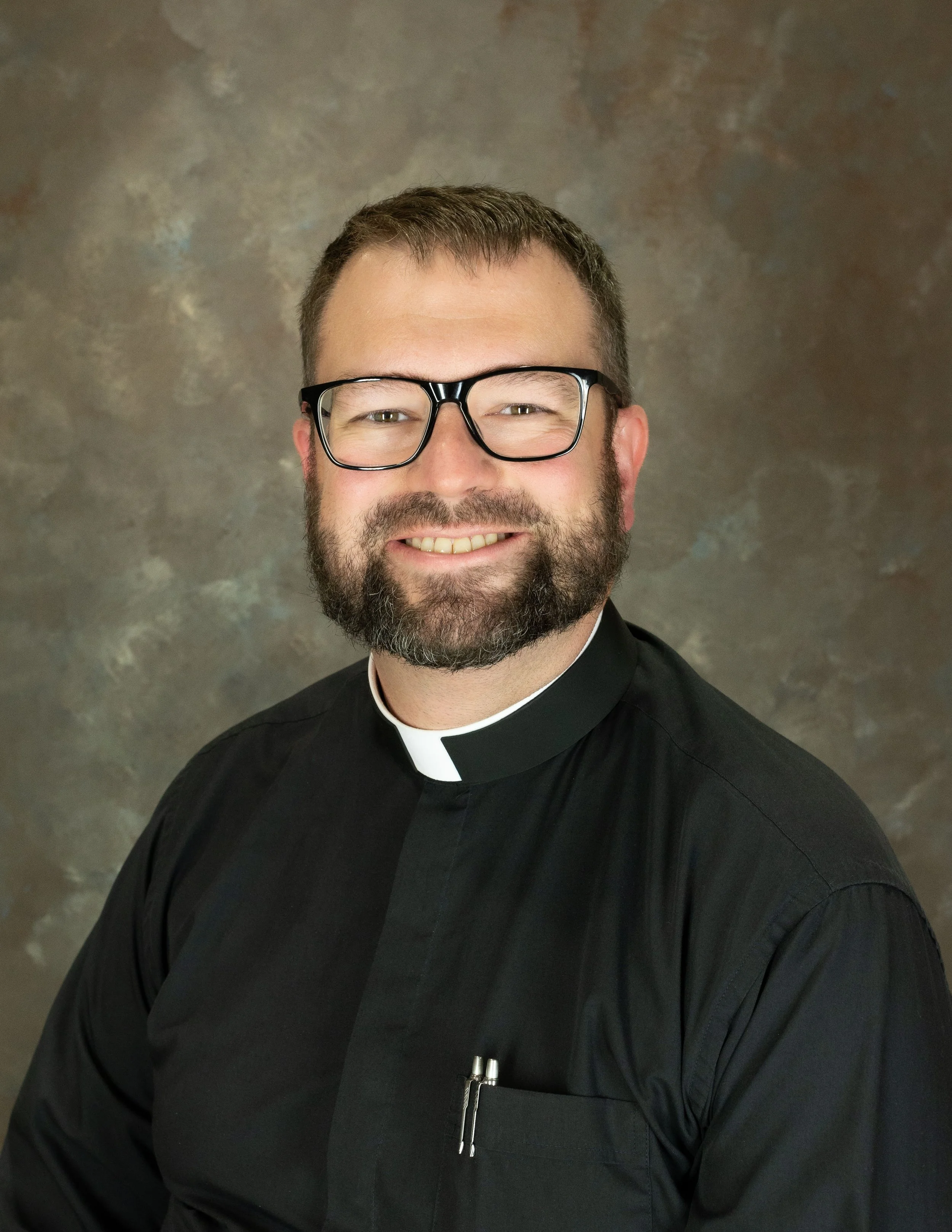 A smiling man wearing glasses and clerical clothing with a white clerical collar, posing against a dark, textured background.