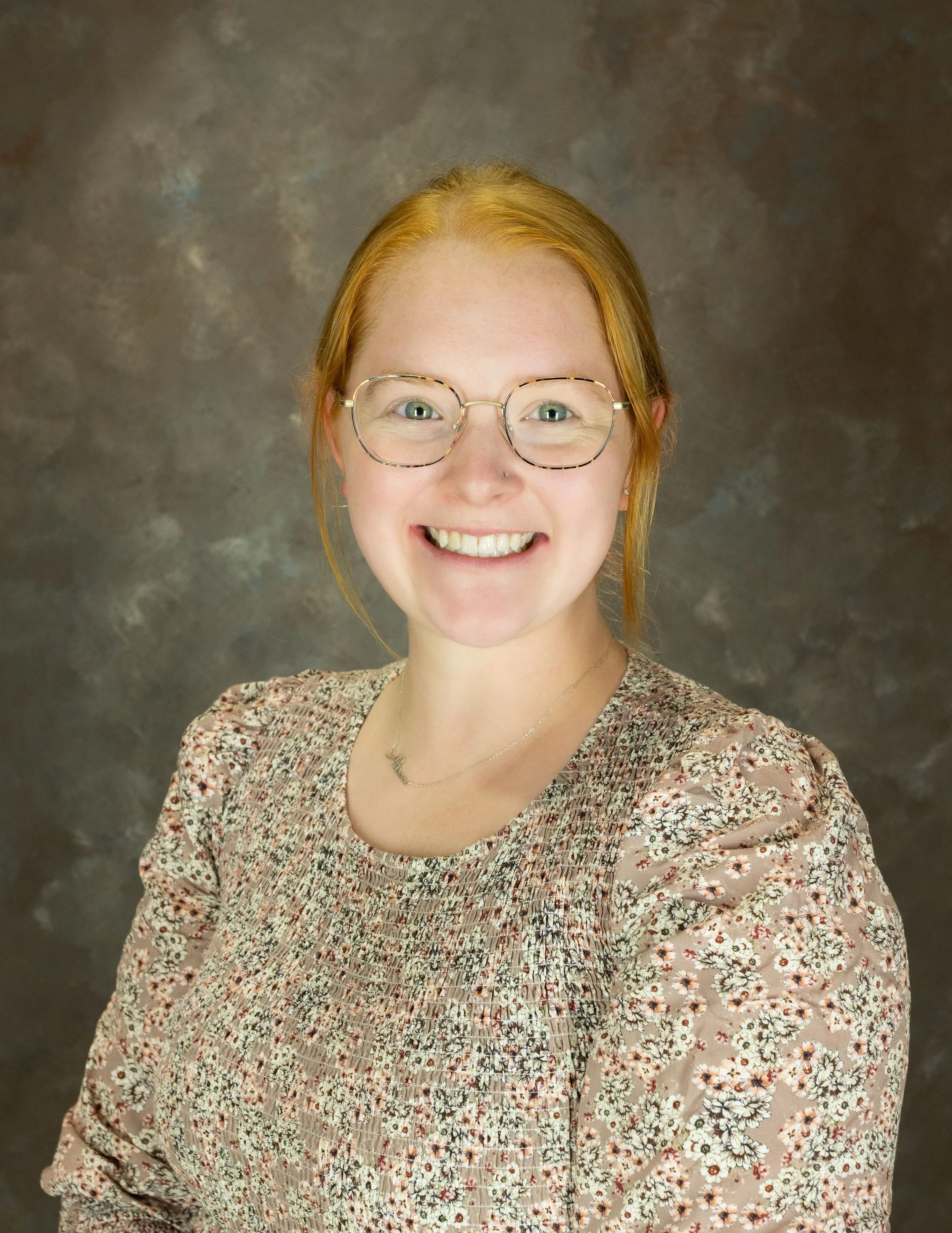 A woman with long red hair smiling at the camera, wearing a green blouse and beige cardigan, against a dark, textured background.