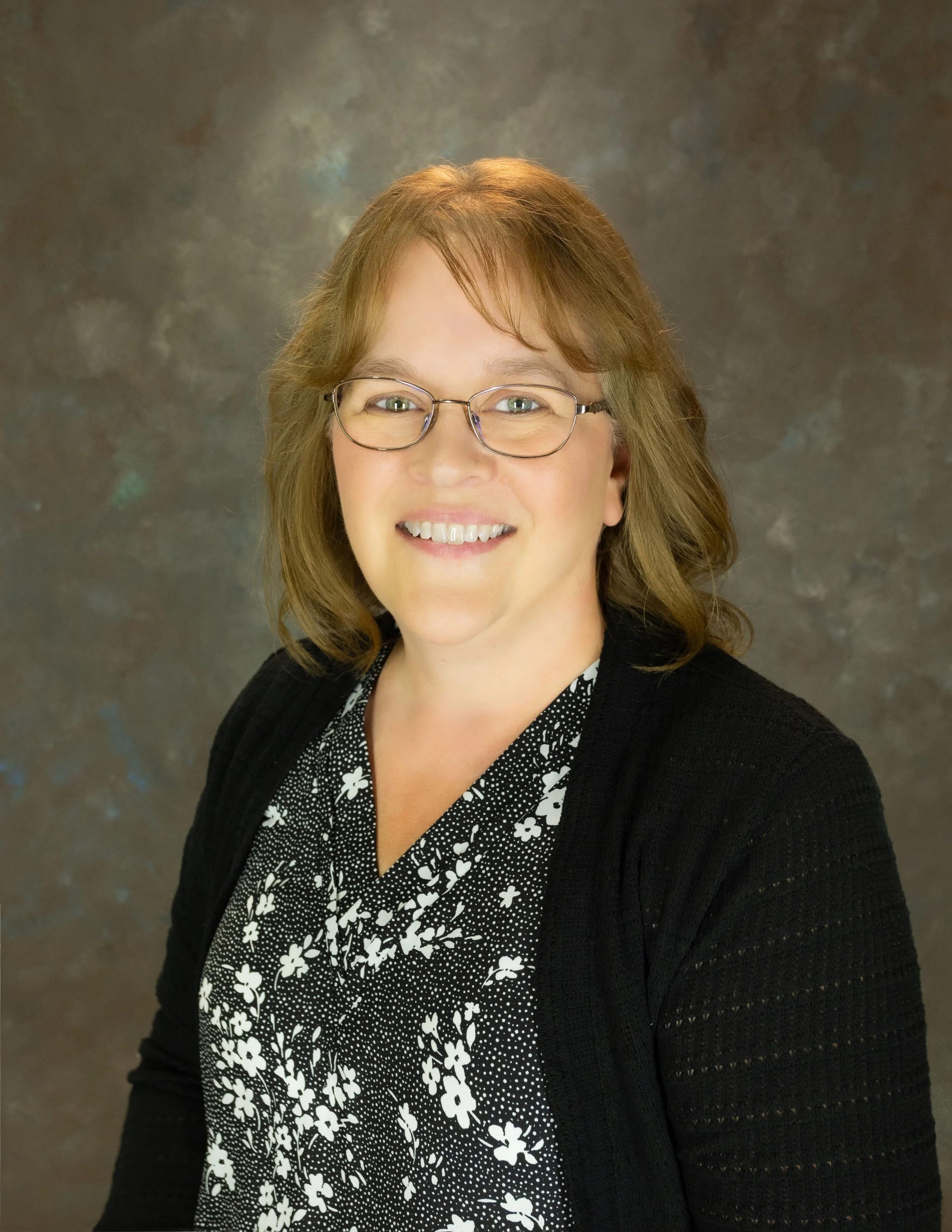 A professional woman with glasses and shoulder-length wavy hair, smiling, wearing a black jacket over a black top with a white floral embroidery, and a beaded necklace, against a dark, textured background.