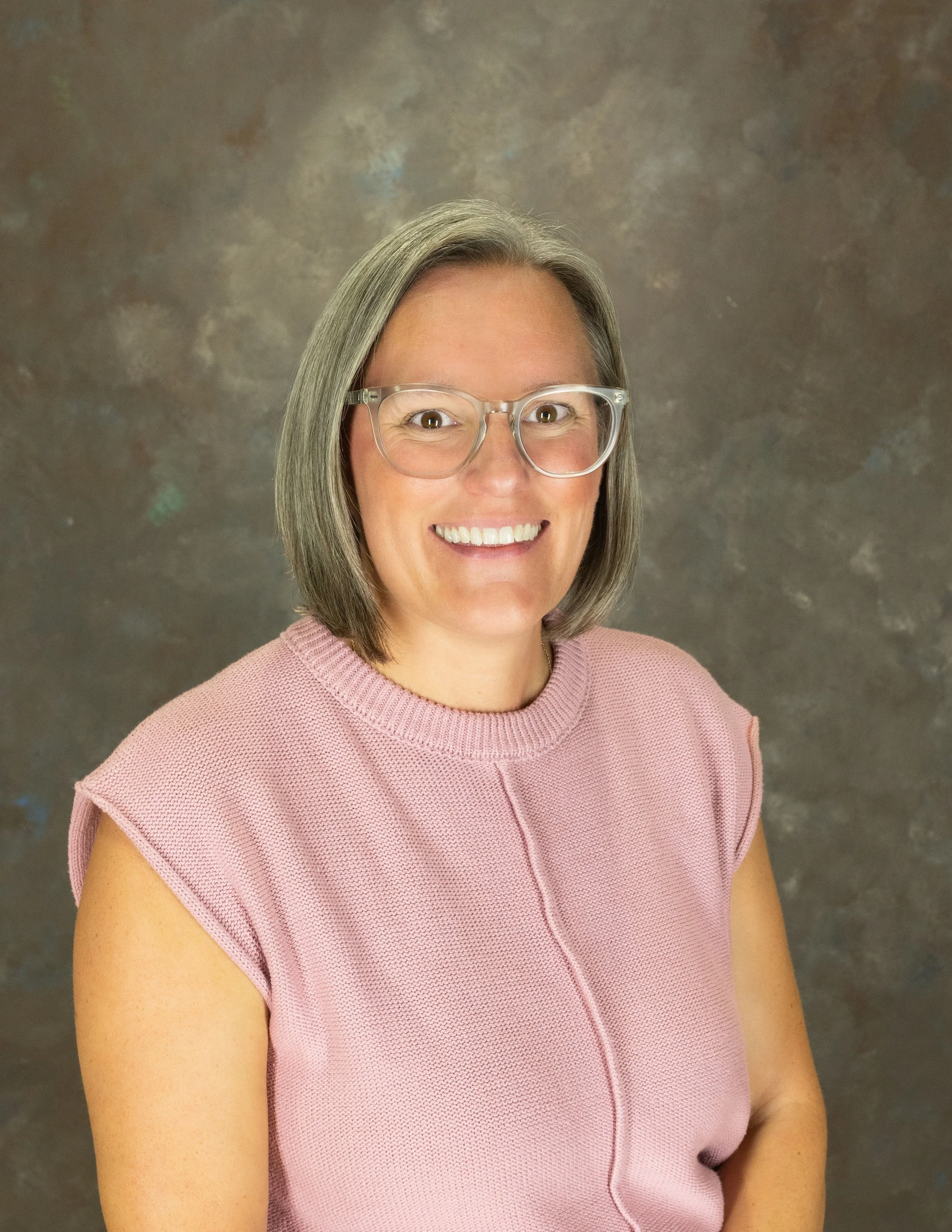Portrait of a woman with short gray hair, wearing glasses, a pink shirt against a brown mottled background.