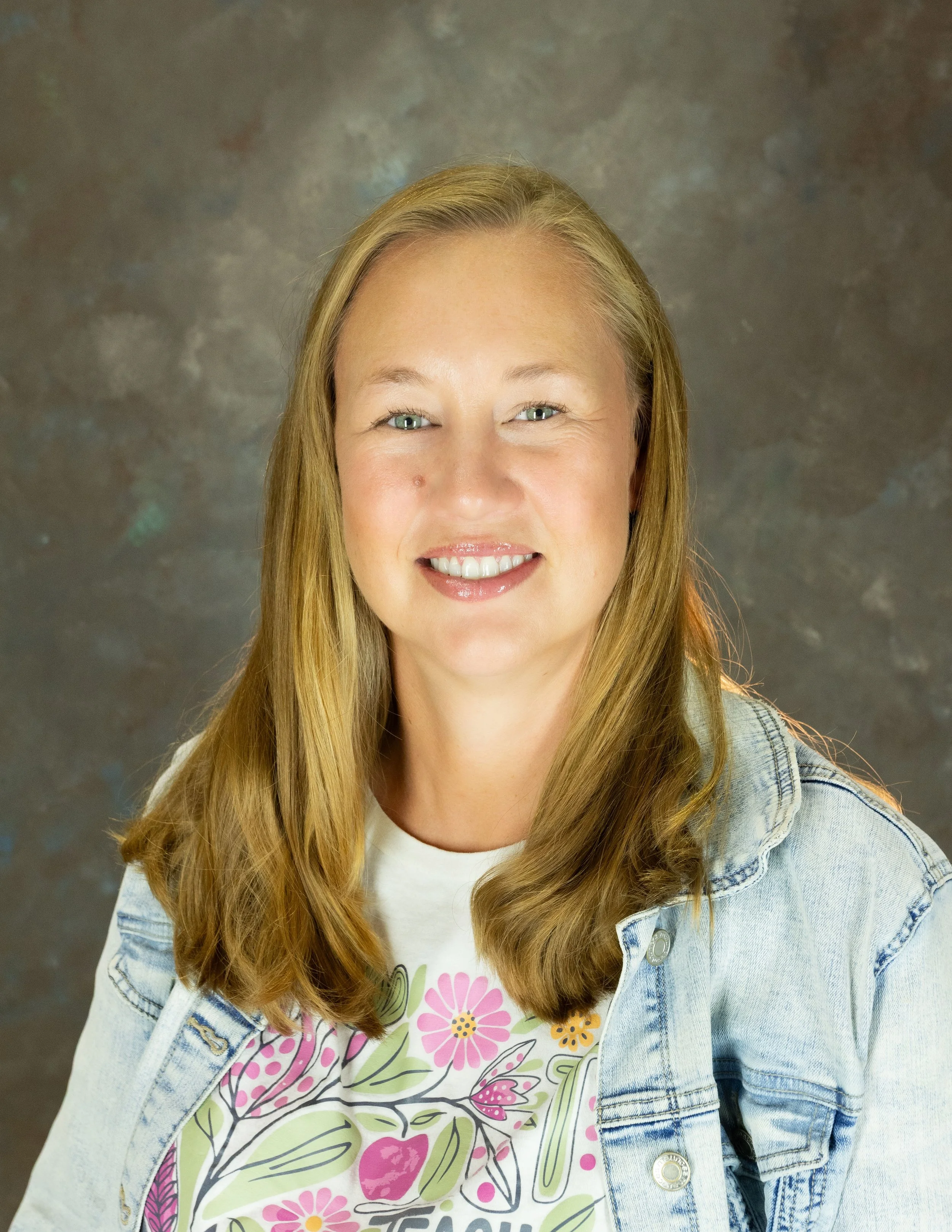 A woman with blonde hair smiling, wearing a gray shirt and purple cardigan against a brown textured background.