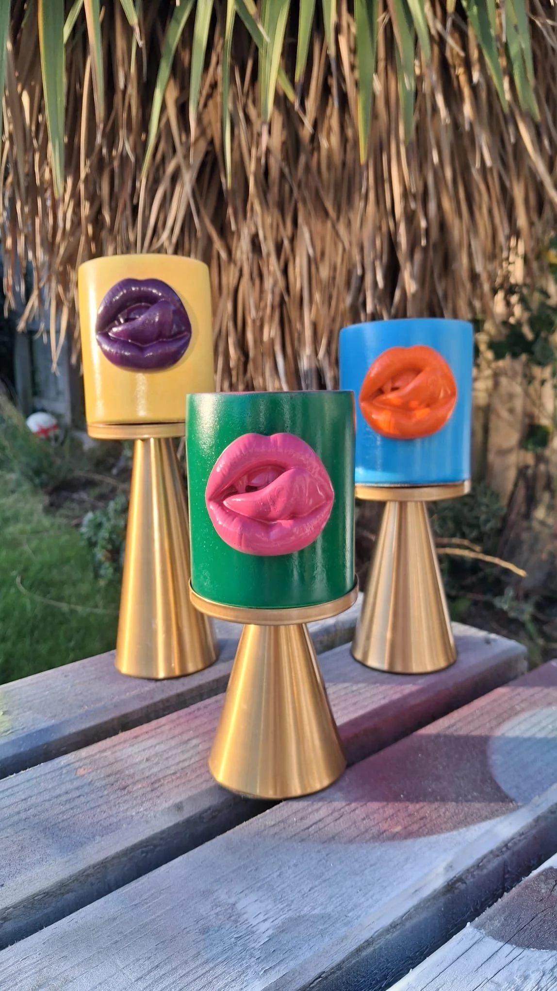 Three candle holders with lips designs in yellow, green, and blue, placed on a wooden surface outdoors with a background of a dried plant or tree.