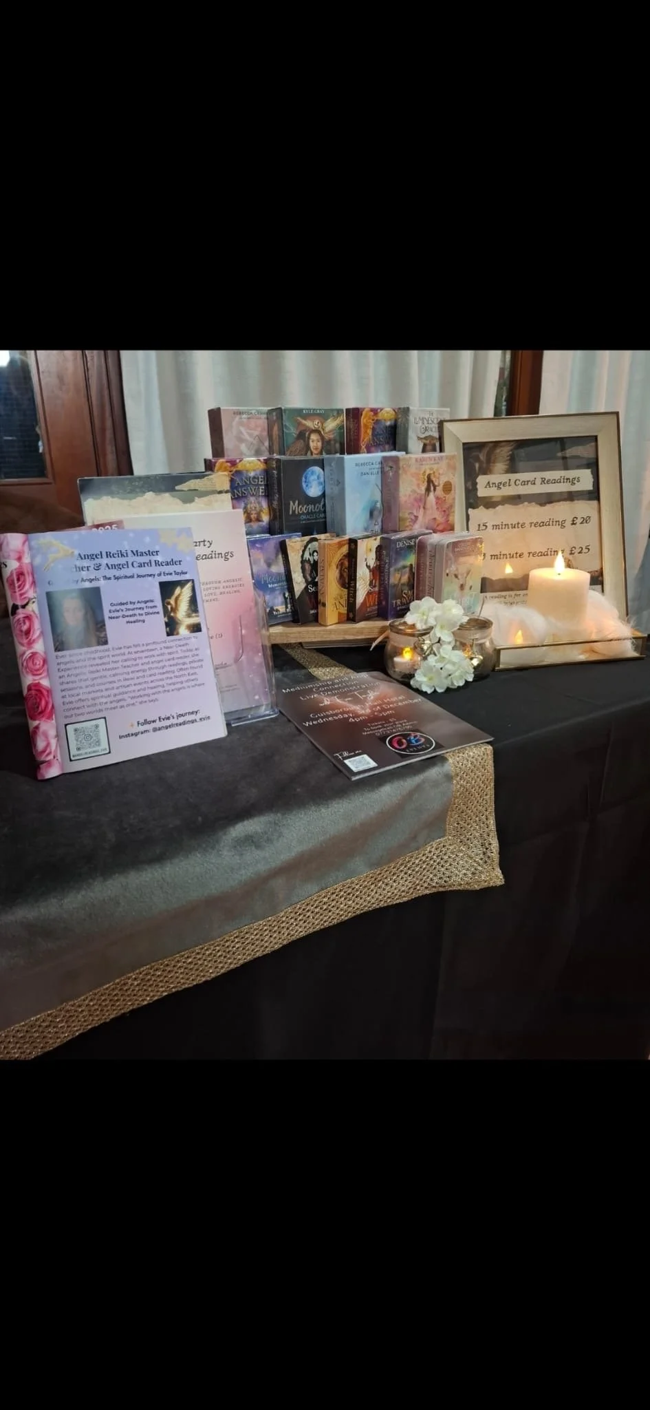 A display table with various tarot and oracle card decks, framed signs for angel card readings, candles, white flowers in glass jars, and informational flyers about angel reiki master and card readings.