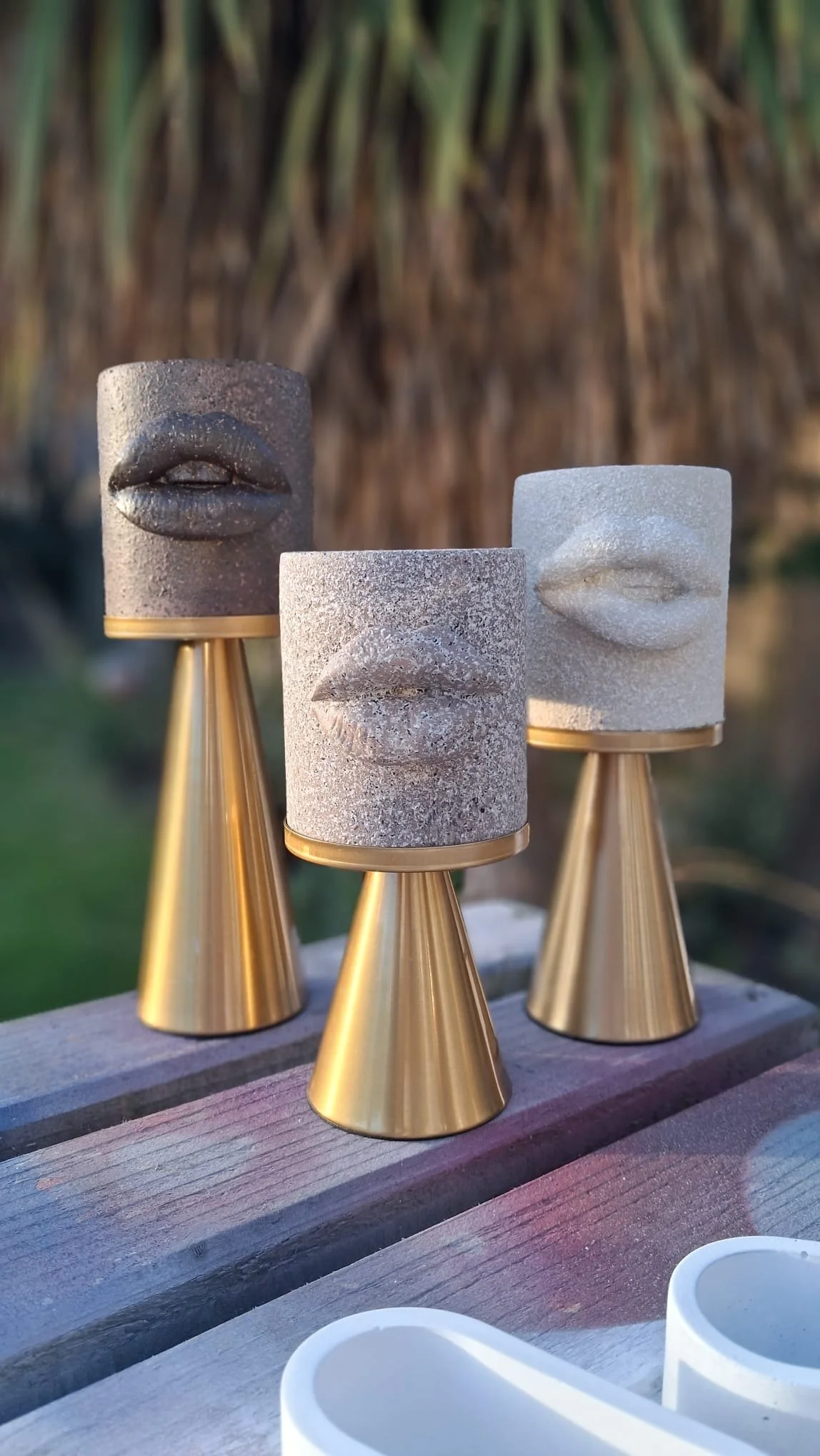 Three decorative sculptures of human lips on cylindrical bases with gold-colored conical stands, placed on a wooden surface outdoors.