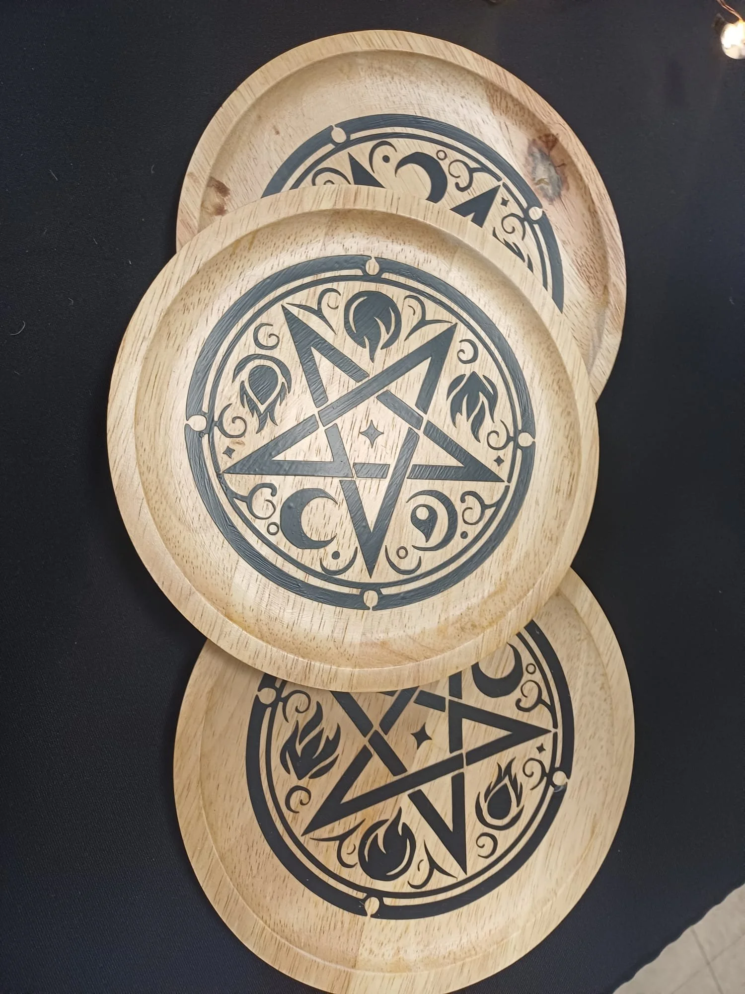 Three wooden coasters with black pentagram and occult symbols printed on their surface, stacked on top of each other on a black surface.