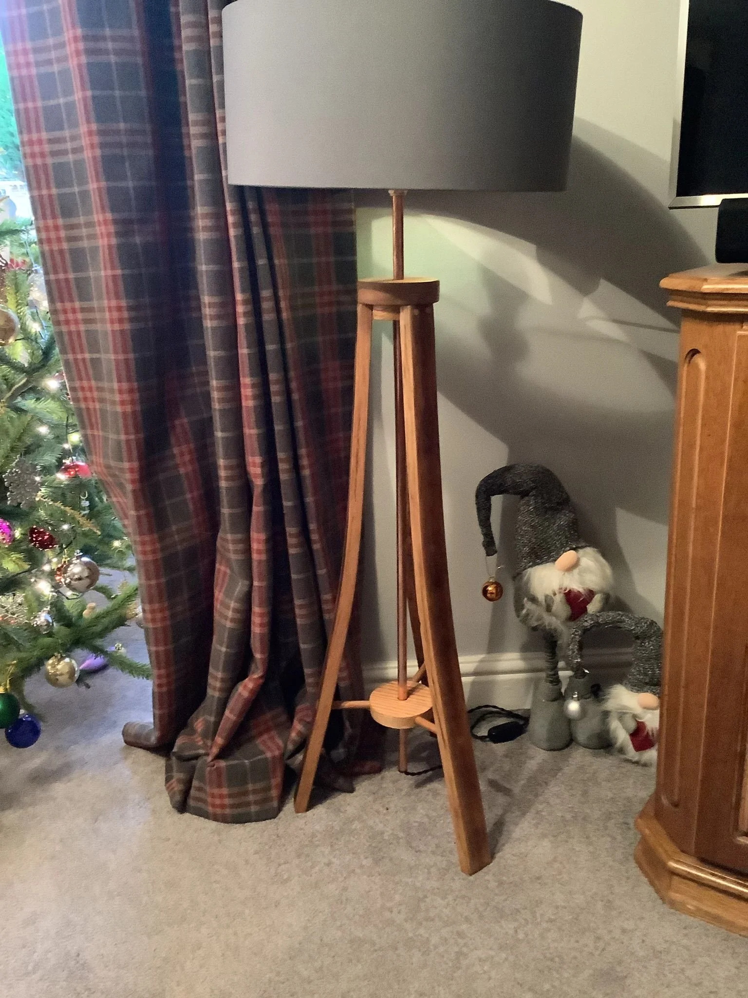 A decorated Christmas tree with ornaments next to a plaid curtain, a wooden floor lamp with a grey shade, and a Santa Claus figurine in a grey hat sitting on the floor near a wooden cabinet.