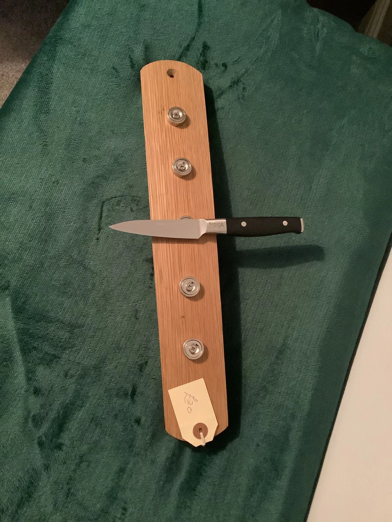 A wooden display board with four metal wheels attached, holding a kitchen knife with a black handle. There is a price tag marked $30 at the bottom of the board. The background is a green fabric surface.