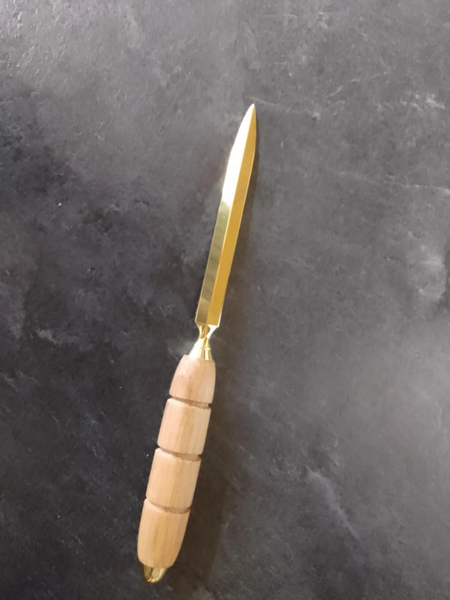 A butter knife with a gold-colored blade and a handle made of three rounded wooden beads, placed on a dark surface.