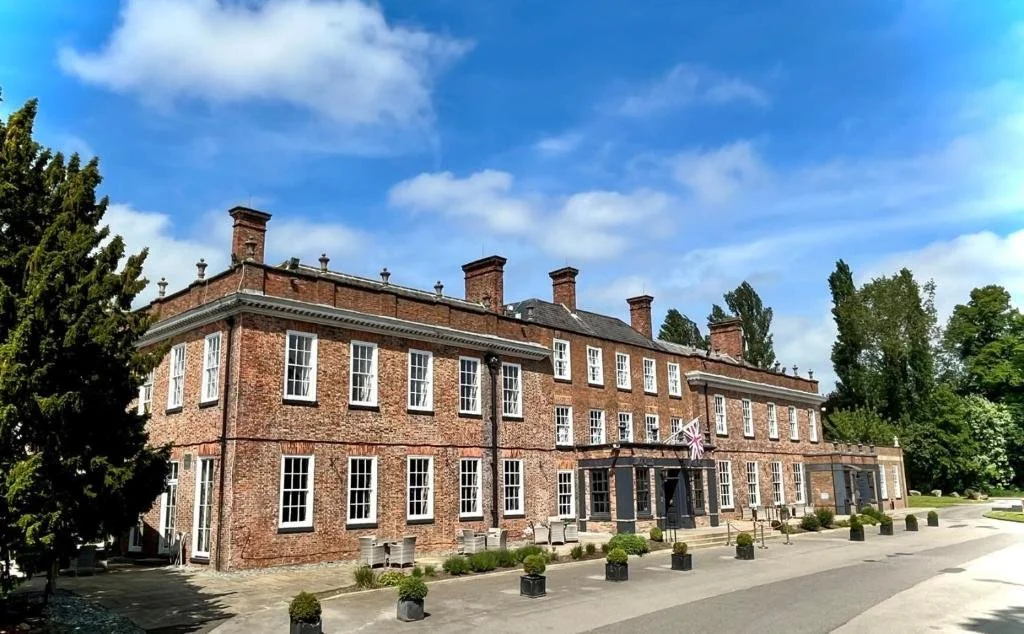 Historic brick building with multiple windows, surrounded by trees, under a partly cloudy sky.