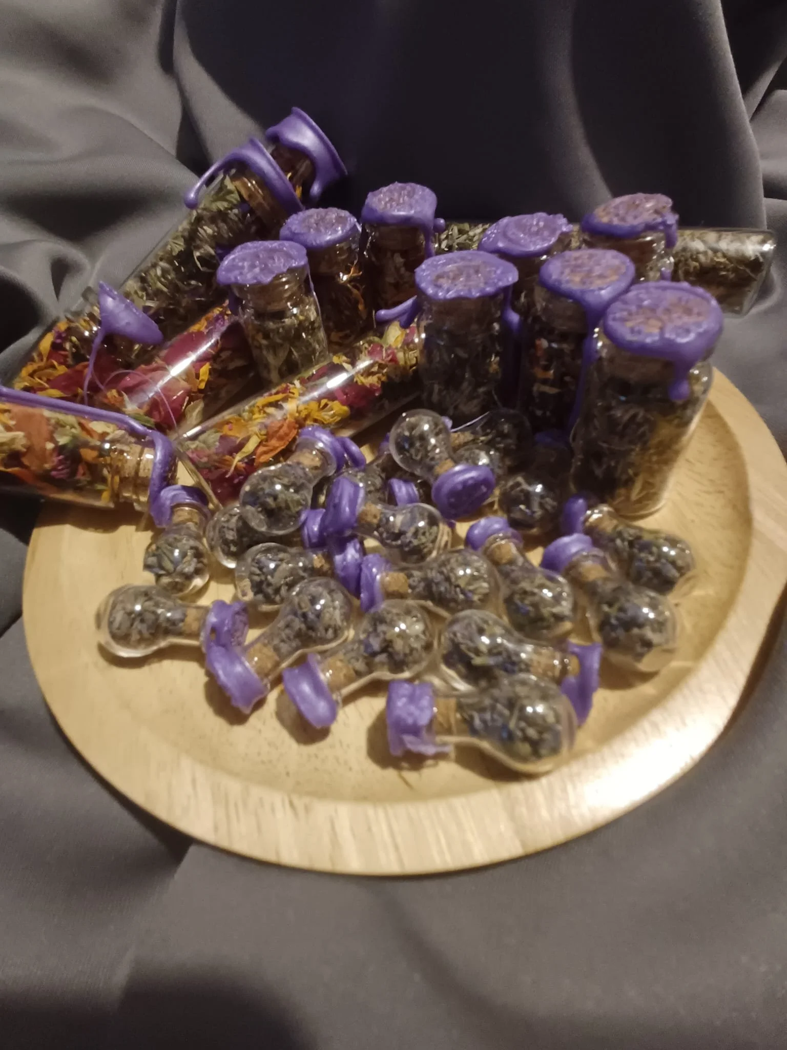 A wooden tray filled with small glass jars with purple lids and some small glass bulbs, all containing dried herbs or flowers.