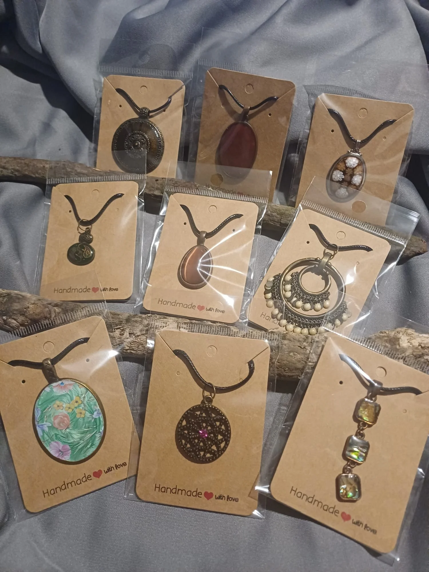 Collection of handmade necklaces with various pendant styles, displayed on cardboard tags. The pendants include a coin, a oval stone, floral design, painted oval, intricate circular design, floral necklace, and square stones, all arranged on a driftwood piece with a gray background.